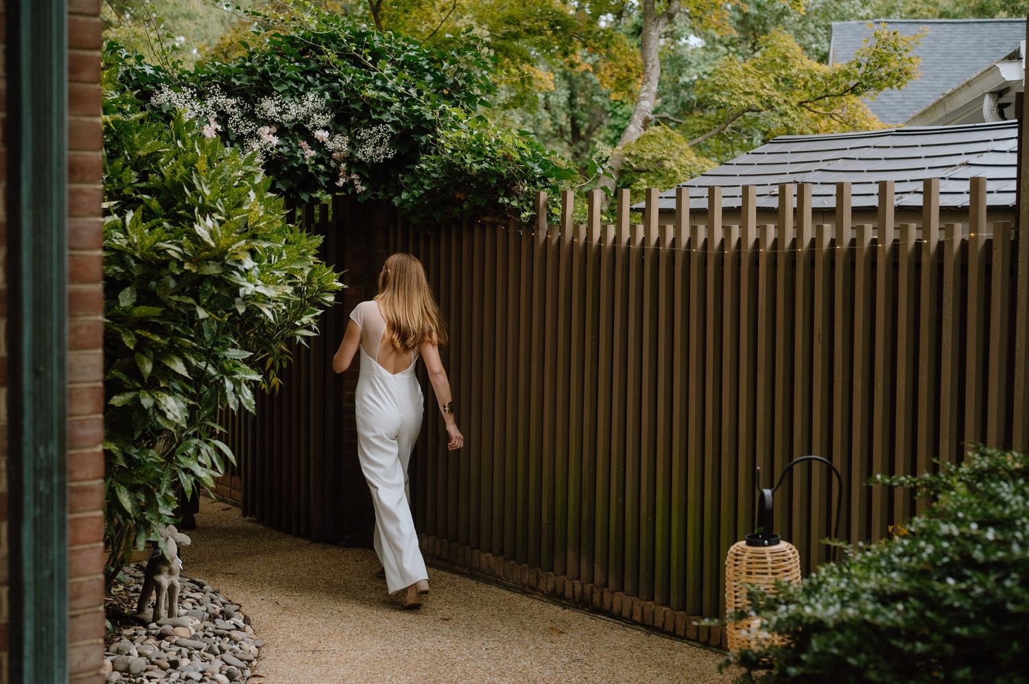 Person in flowing white dress walking along garden path by wooden fence.