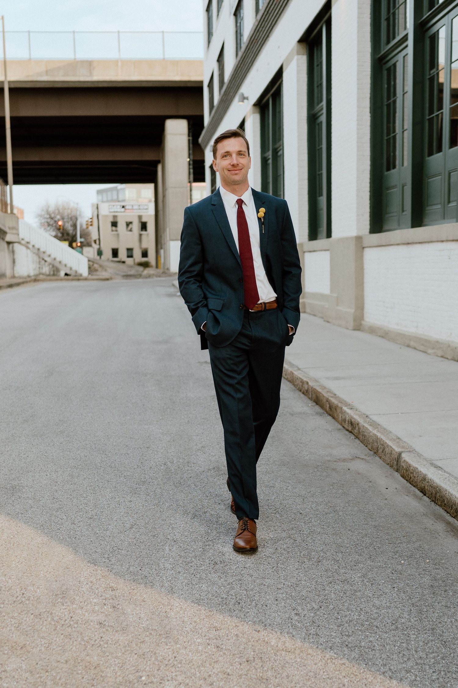 Groom in dark suit and red tie poses confidently on urban street.