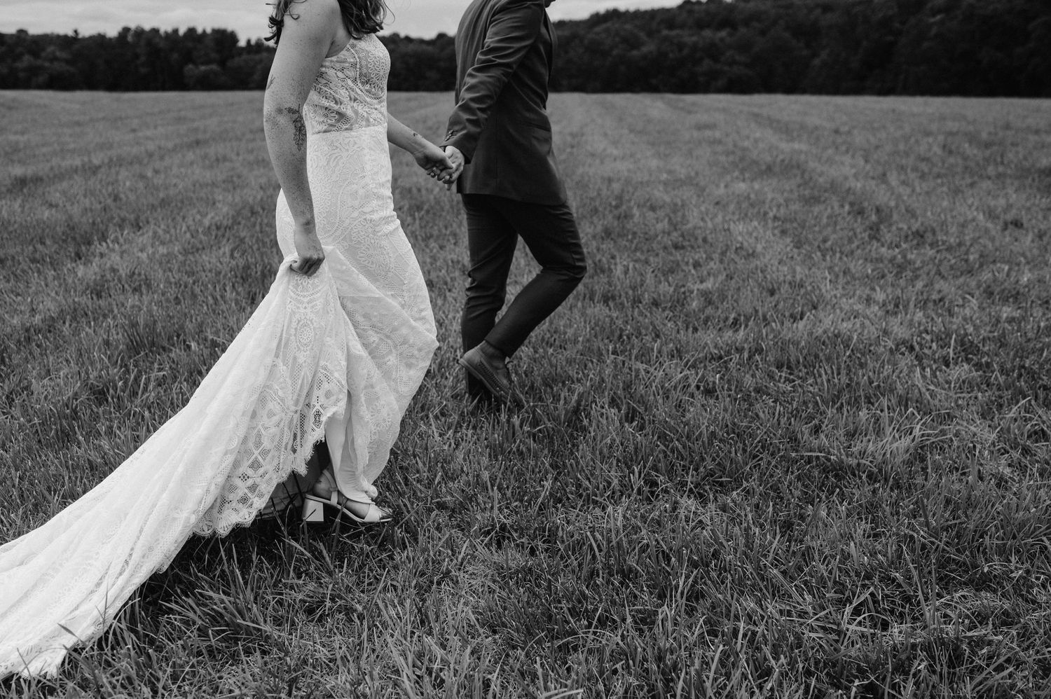 Black and white photo of wedding couple walking through field with trailing dress.