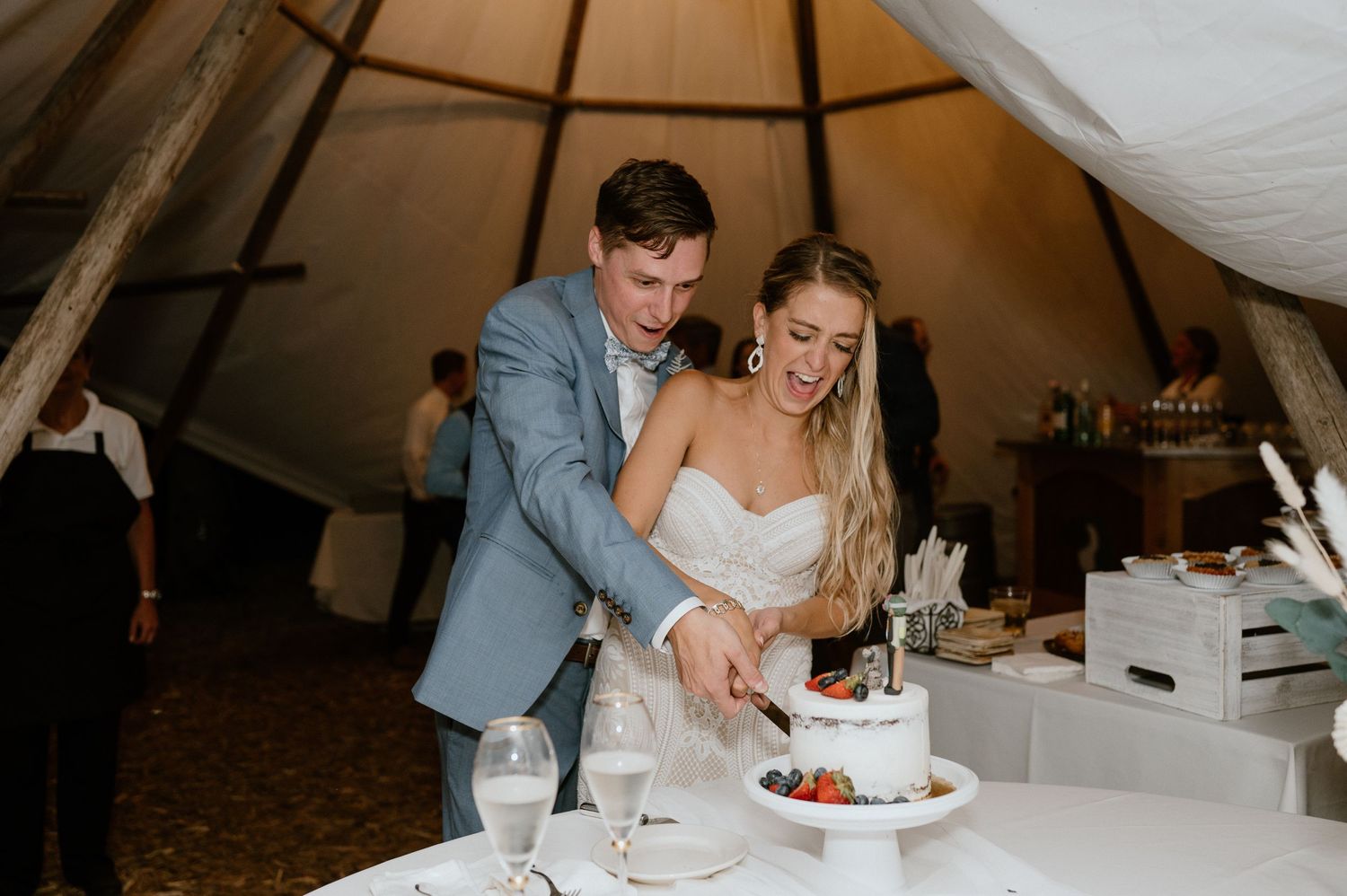 A couple cuts their wedding cake together inside a warmly lit reception tent.