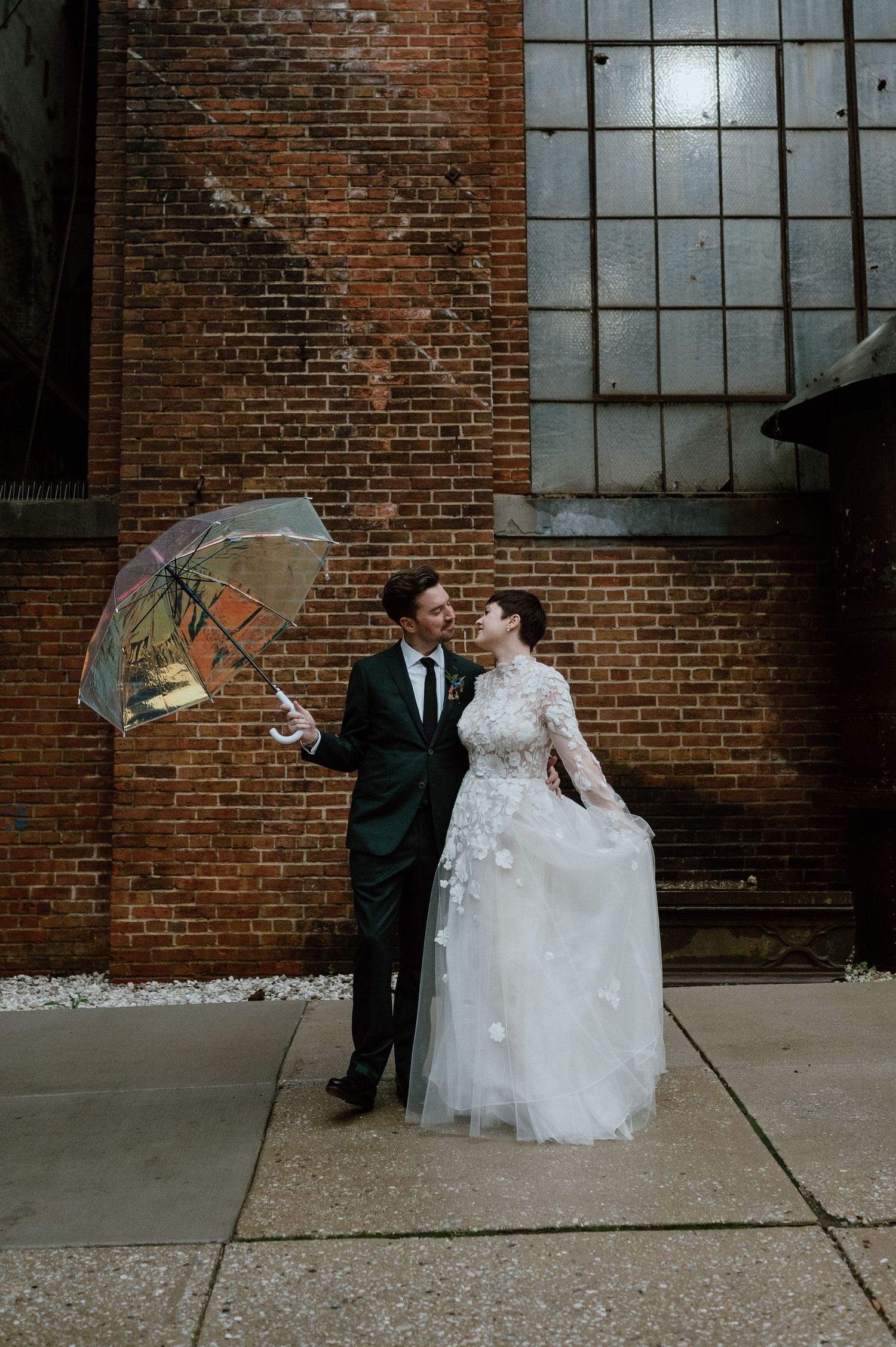 Wedding couple poses with a clear umbrella against brick wall background in urban setting.
