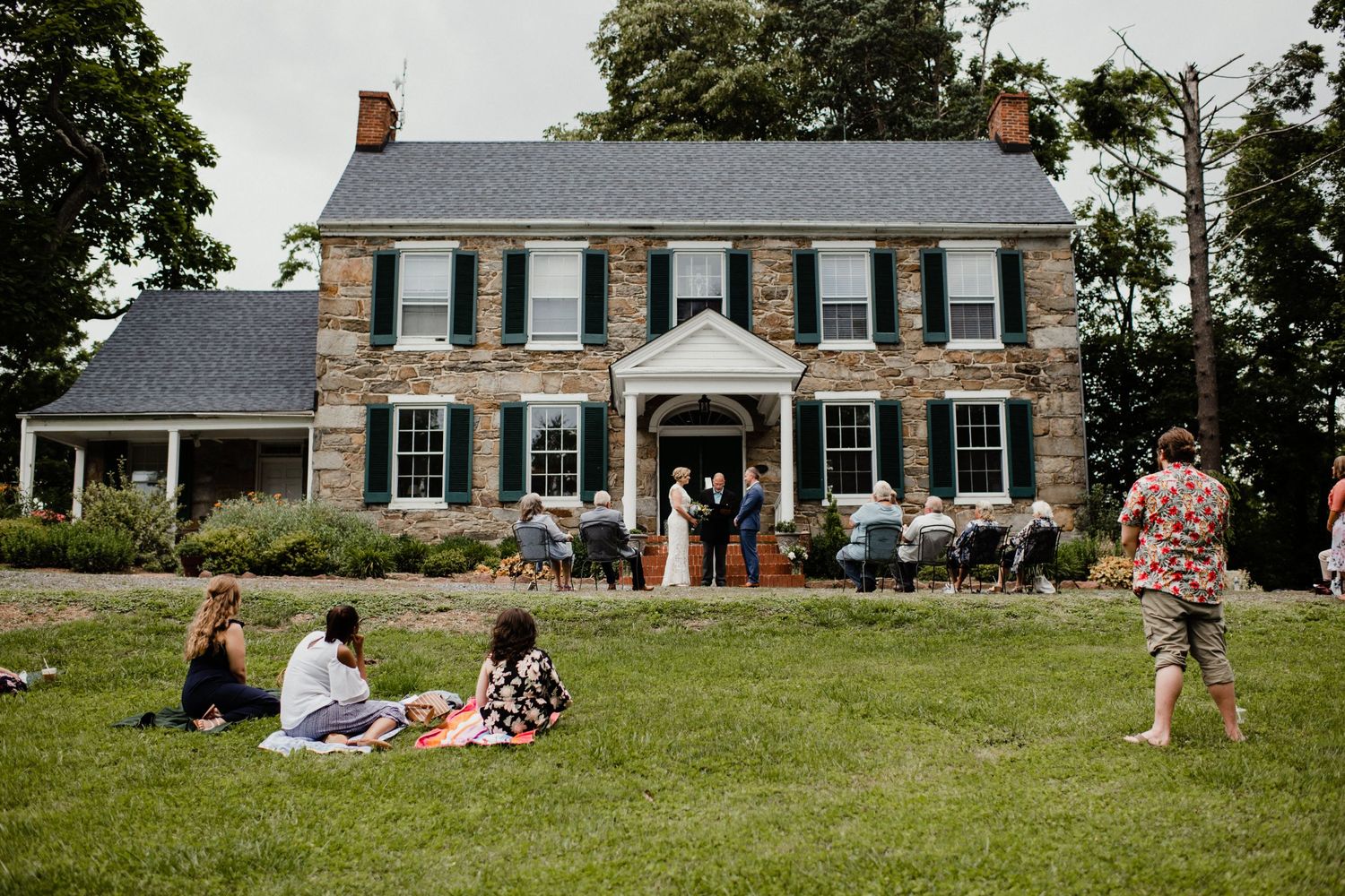 Historic colonial stone house with wedding ceremony and guests on front lawn.