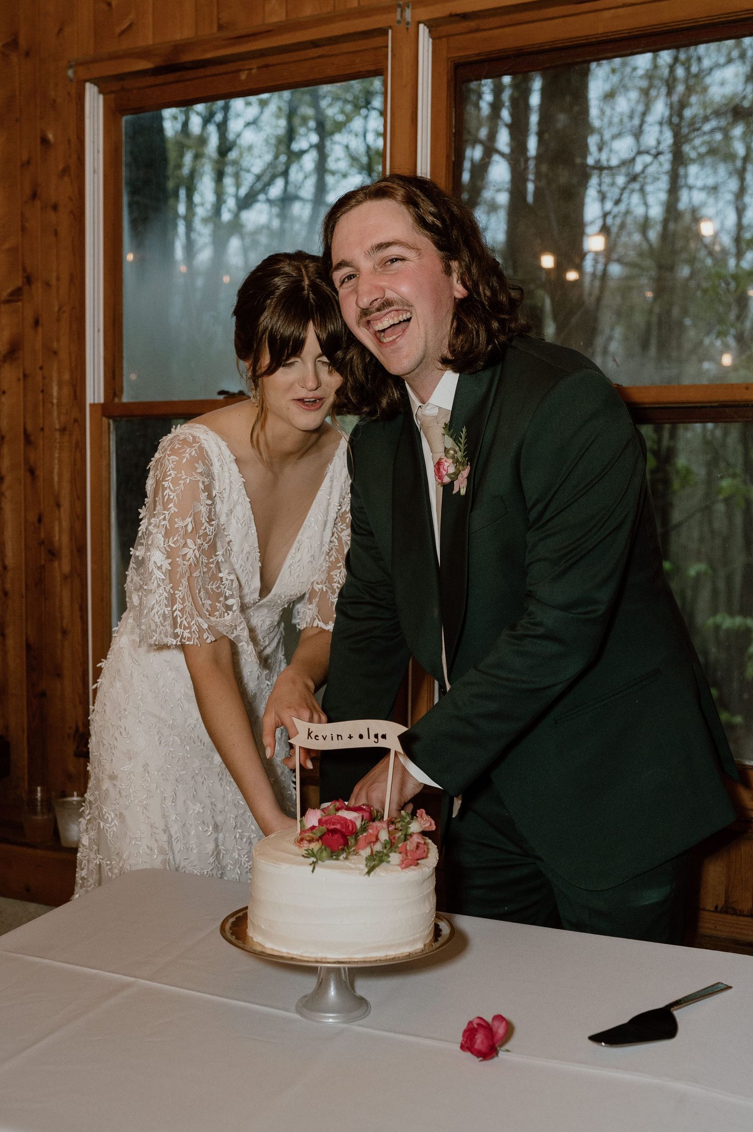 Newlywed couple cutting their white wedding cake together at a rustic venue with windows overlooking trees.