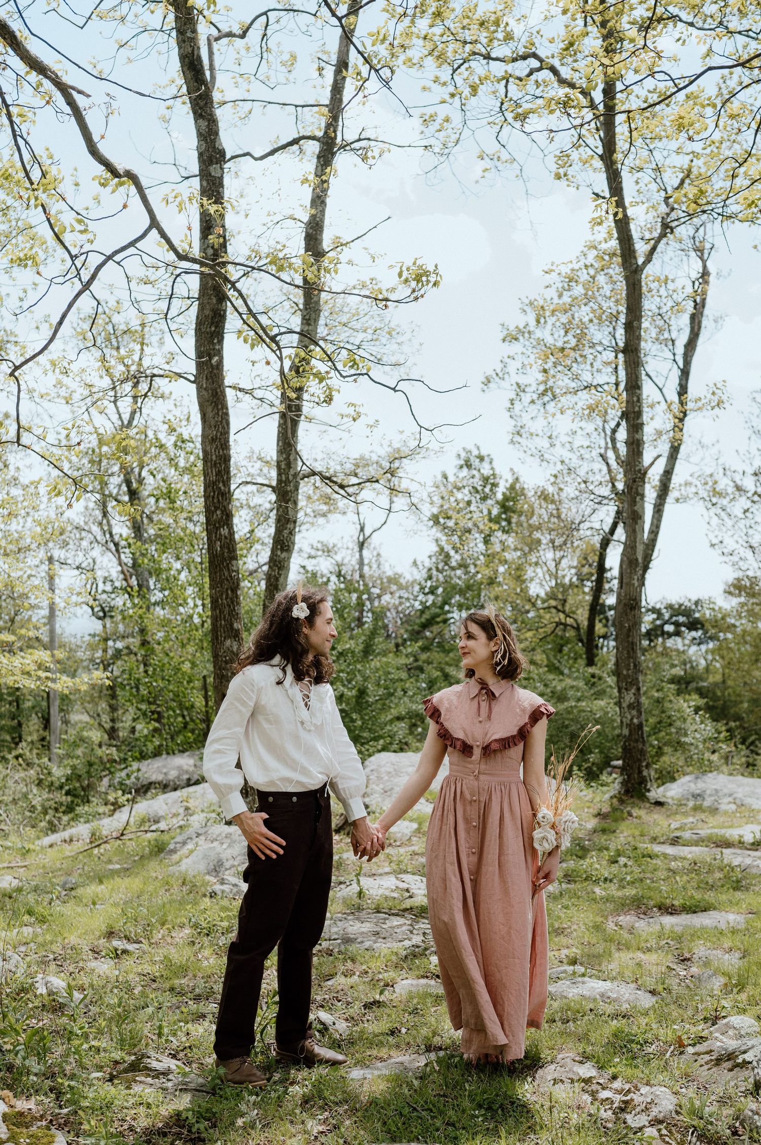 A couple in vintage-inspired clothing stands together in a sunlit forest clearing.