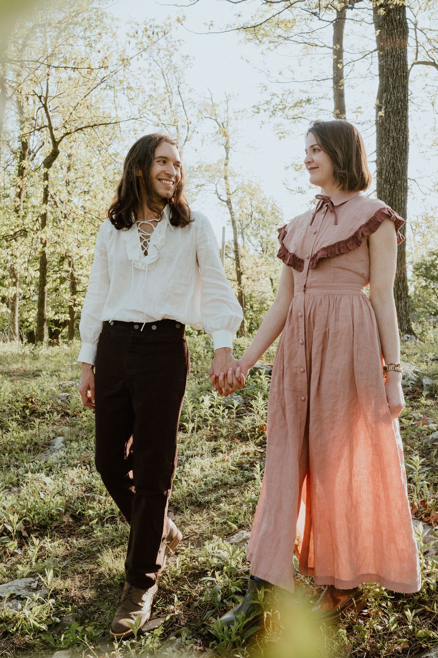 Two people in vintage-inspired outfits share a moment in a sunlit forest setting.