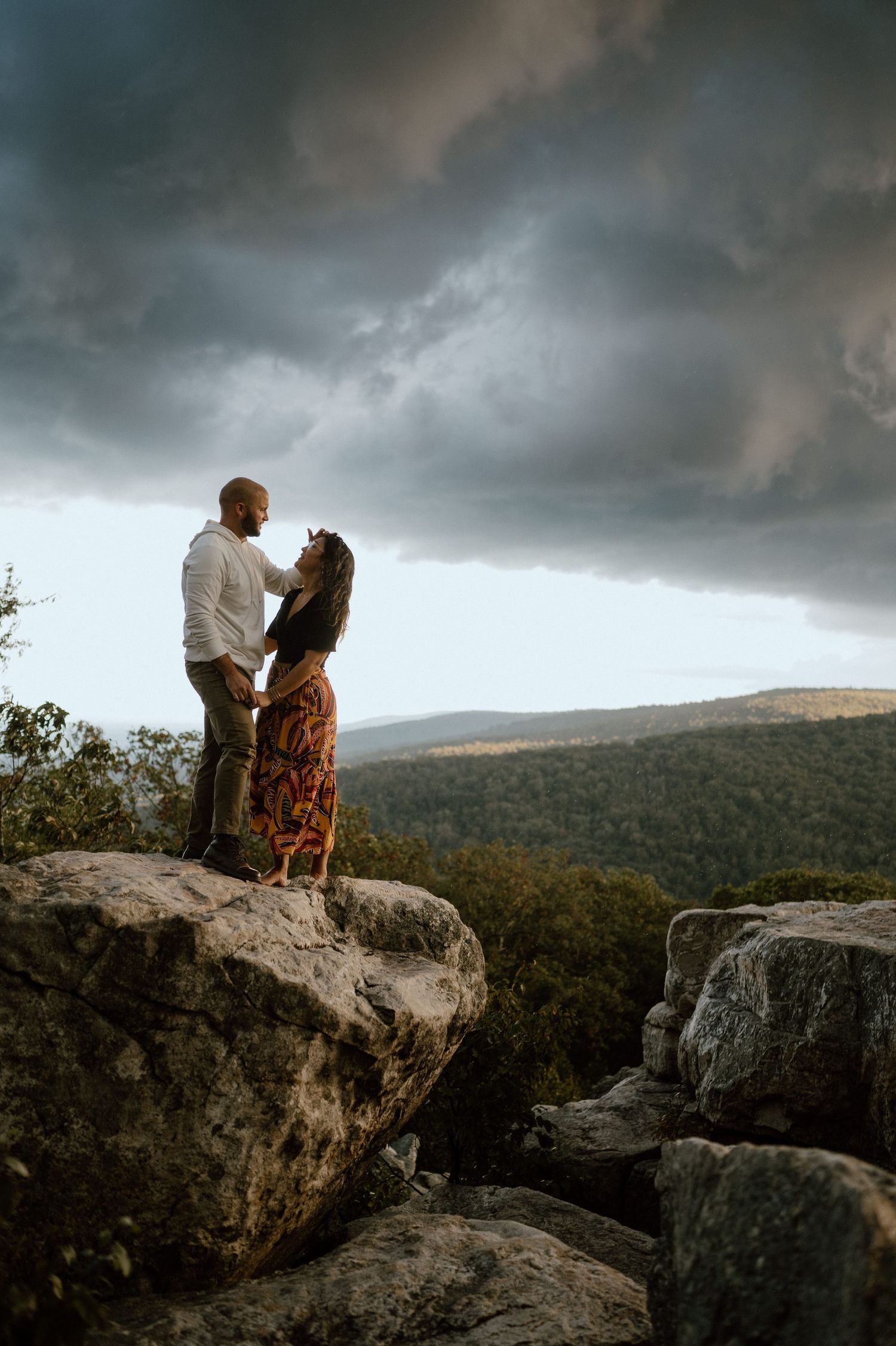 A couple shares an intimate moment on mountain rocks with dramatic storm clouds overhead.