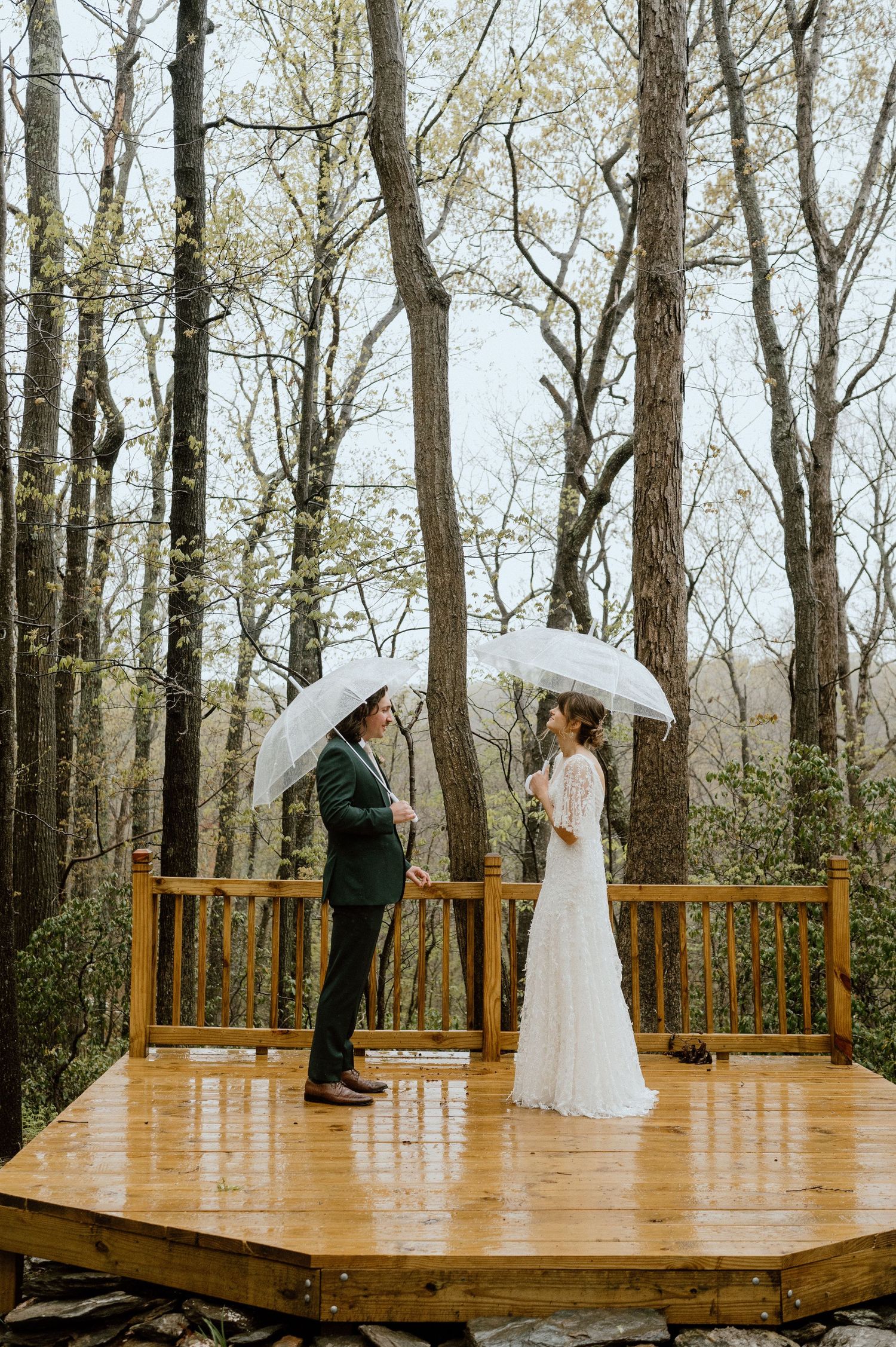 Couple stands facing each other with umbrellas on wooden platform in misty forest.