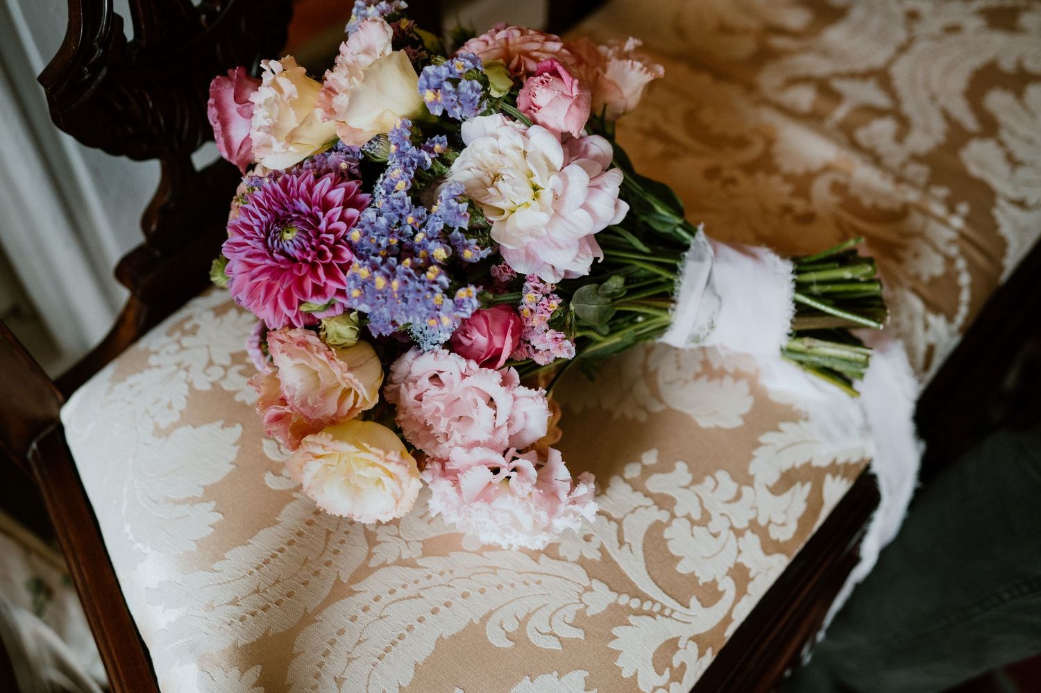 Bridal bouquet with pink peonies and purple flowers resting on damask fabric.