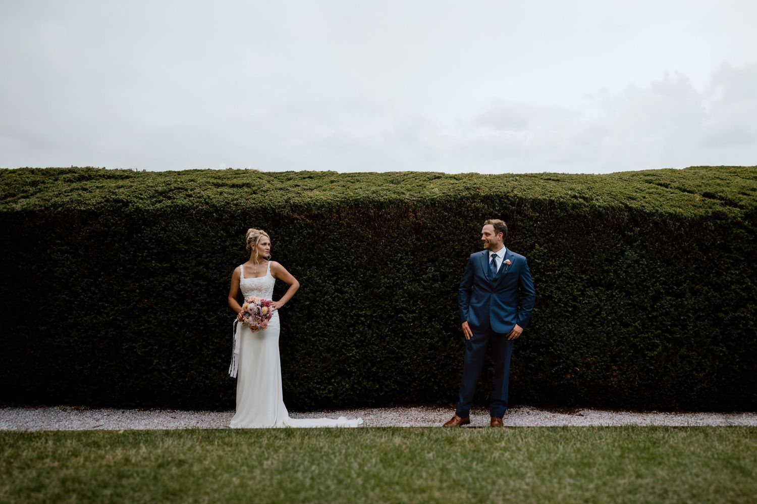 A bride and groom strike playful poses against a formal garden hedge backdrop.