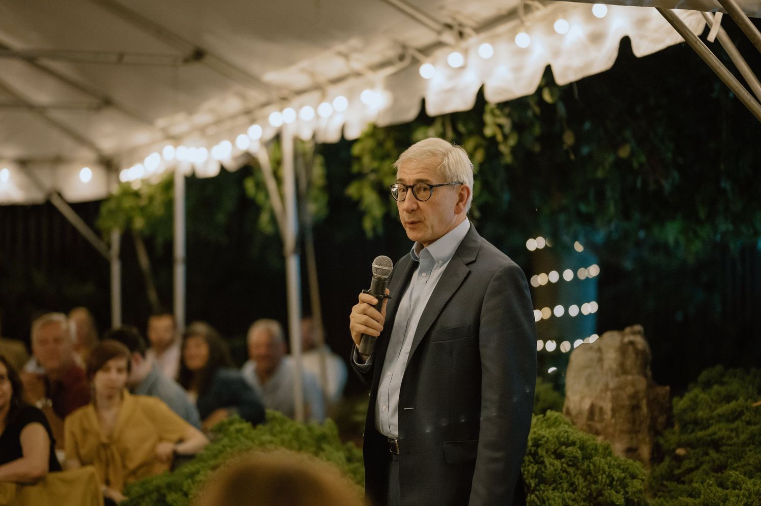Someone giving a speech under tent lighting at an outdoor evening reception.