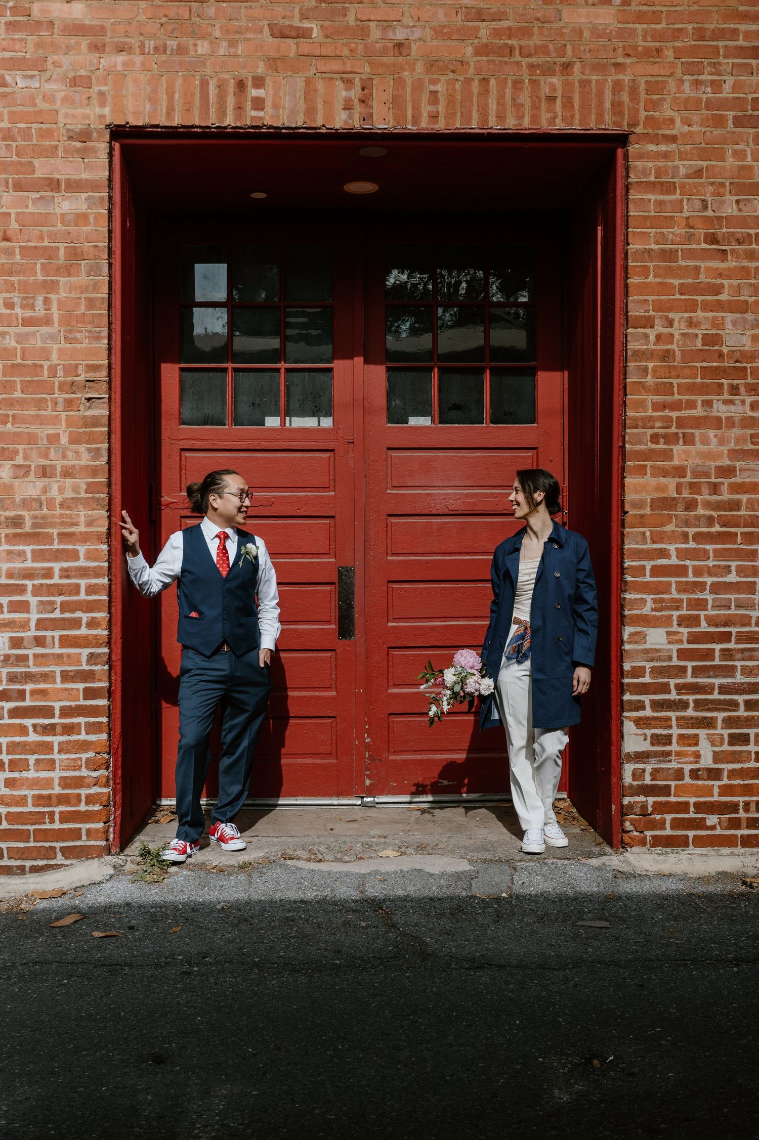 Two people in formal wear stand against red doors of a brick building having a conversation.
