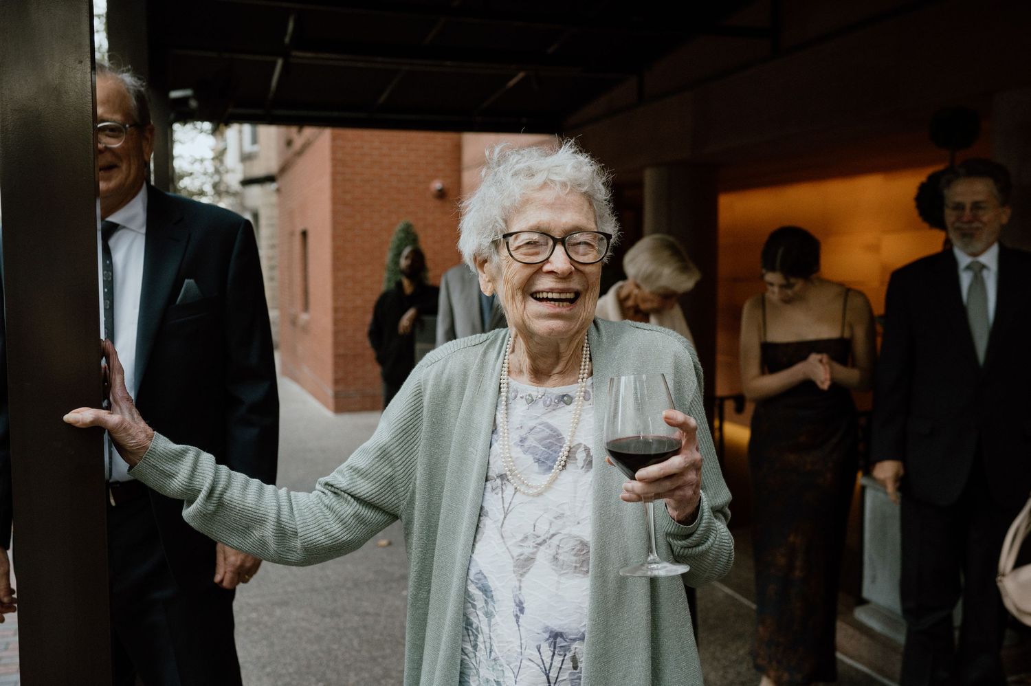 A joyful guest wearing glasses and a light colored outfit smiles while holding a drink at a celebration event.