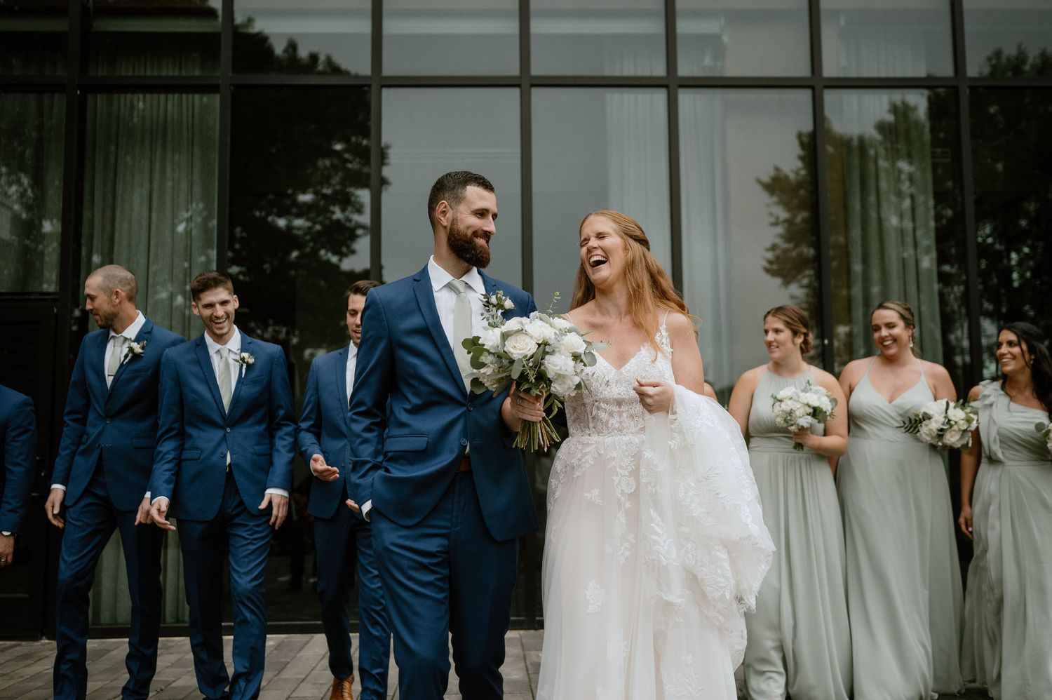 Wedding party poses outside modern glass building with couple in center.