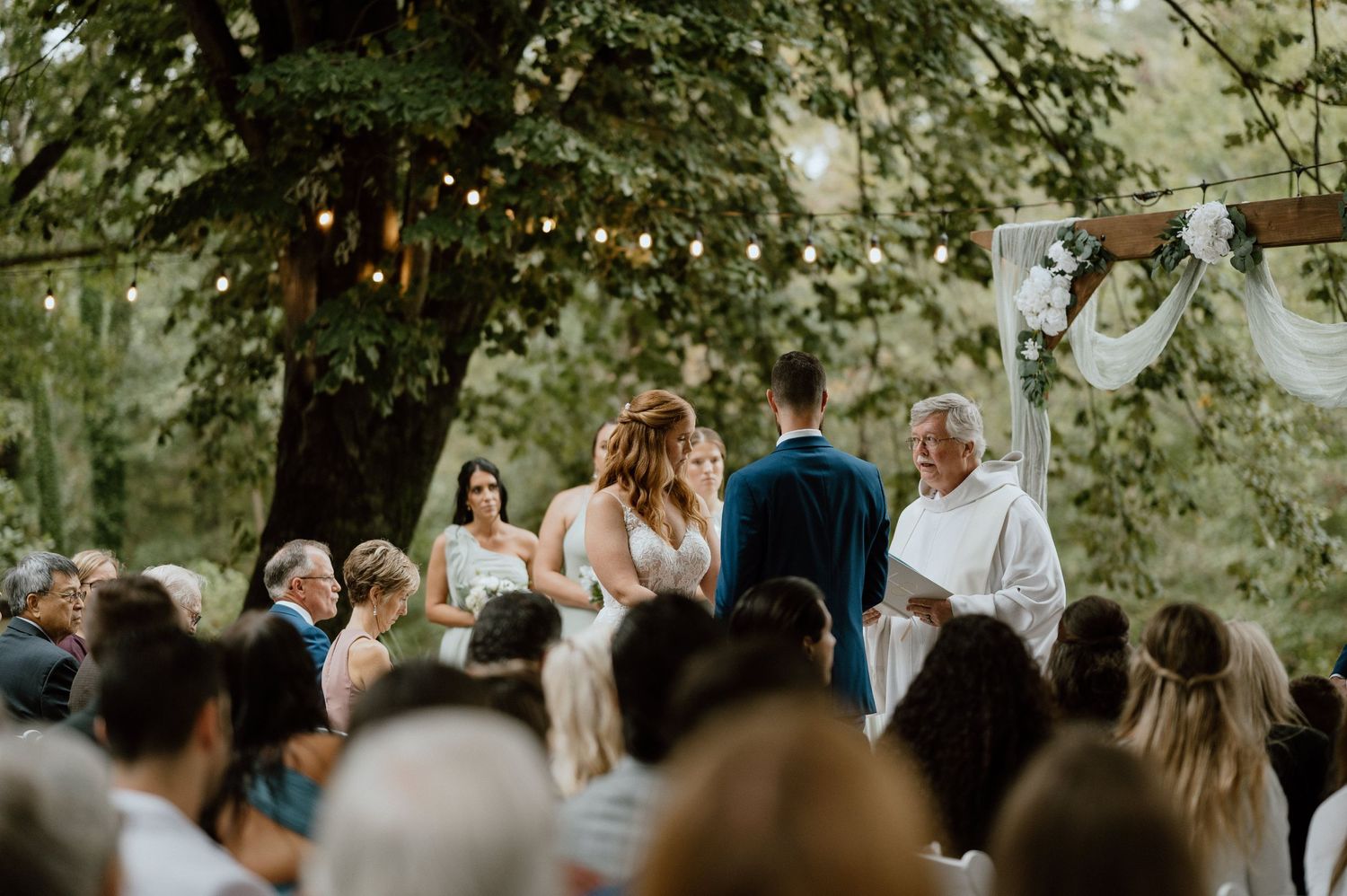 Outdoor wedding ceremony under trees with string lights and gathered guests.