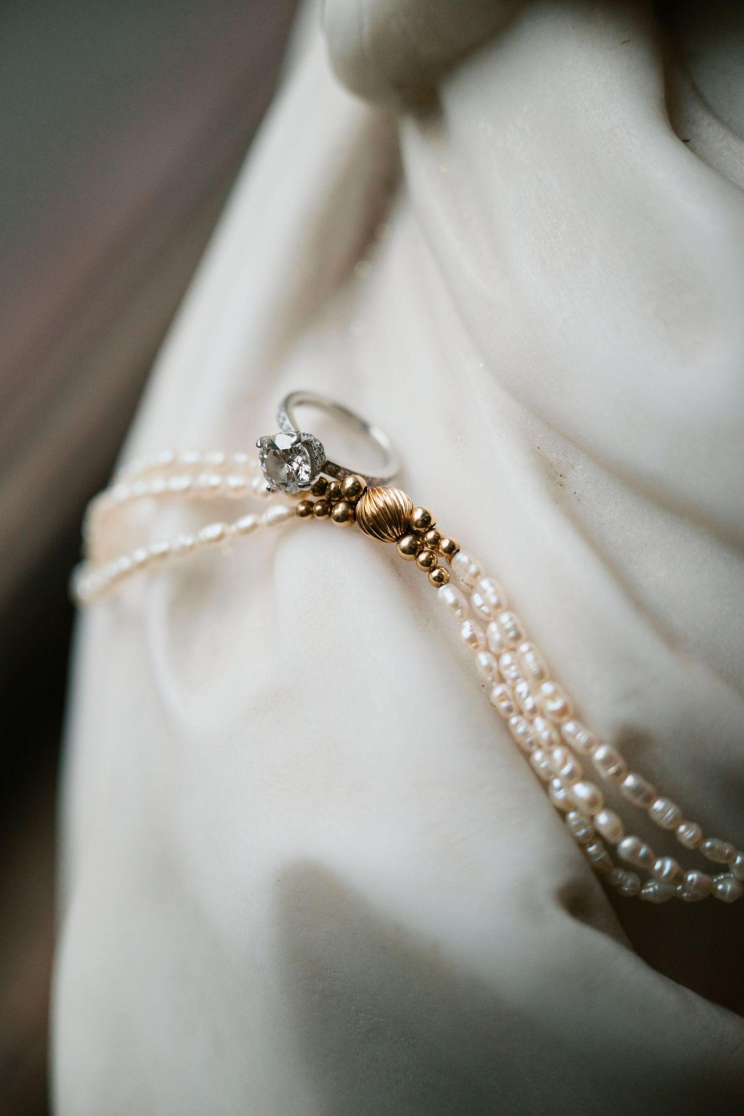 A delicate diamond engagement ring rests on a strand of pearl necklace against white fabric.
