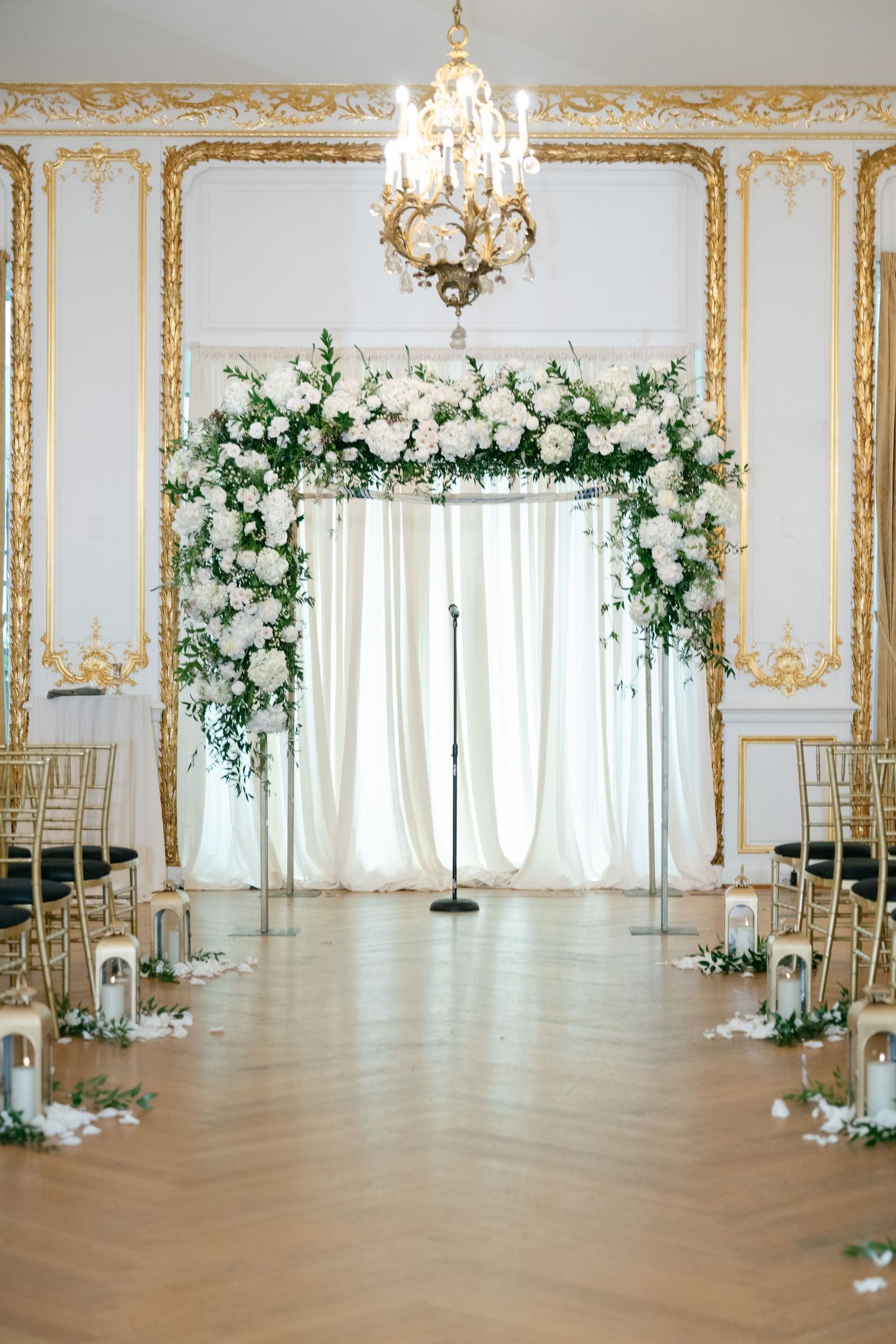 Elegant ballroom with ornate gold trim walls decorated with white floral arrangements and chiavari chairs for a wedding ceremony.