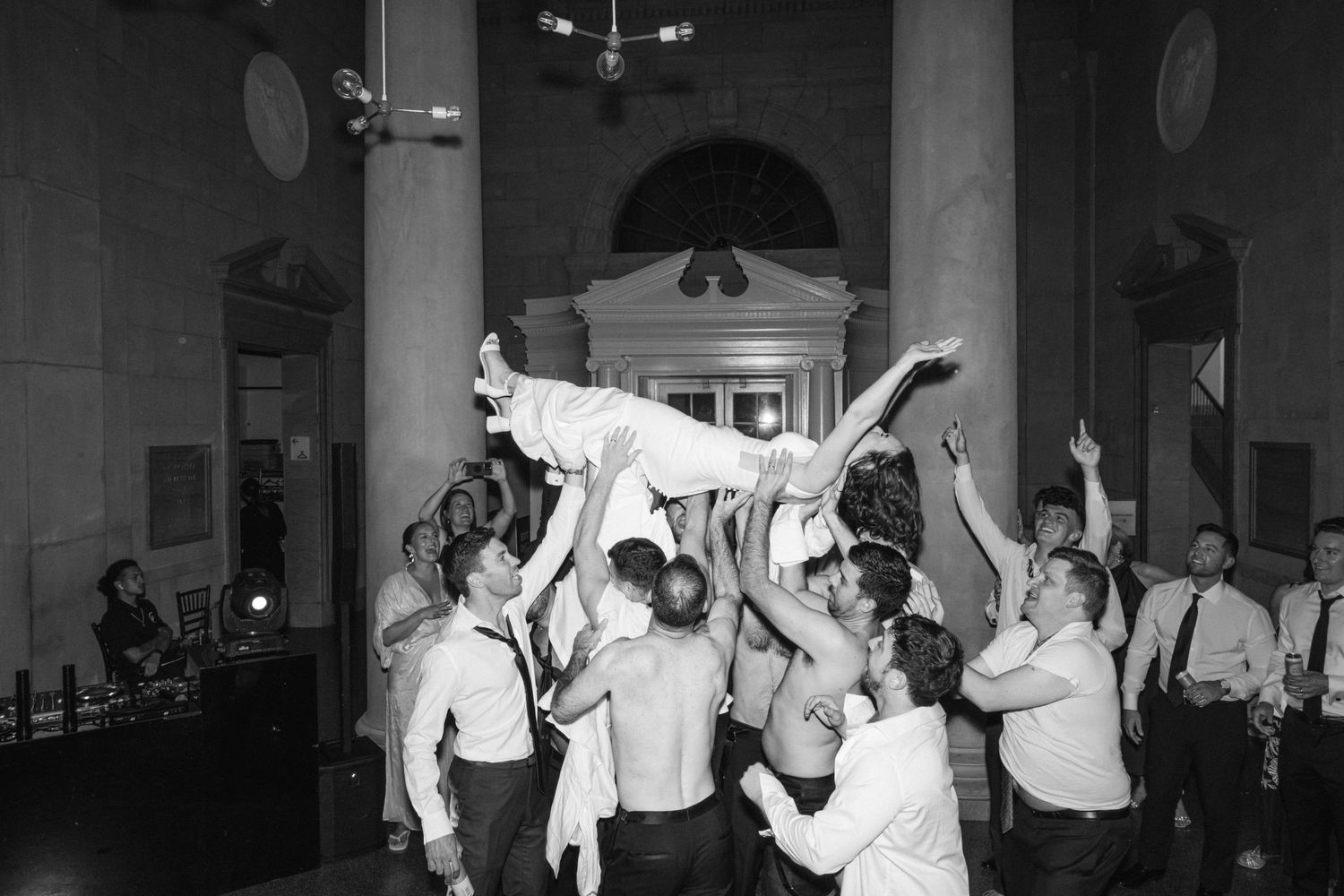 Wedding guests dance and celebrate together at an indoor reception venue in black and white photography.