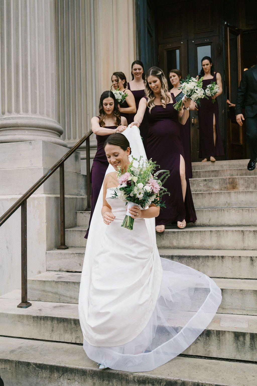 Wedding party in purple dresses watches bride descend grand stone steps.