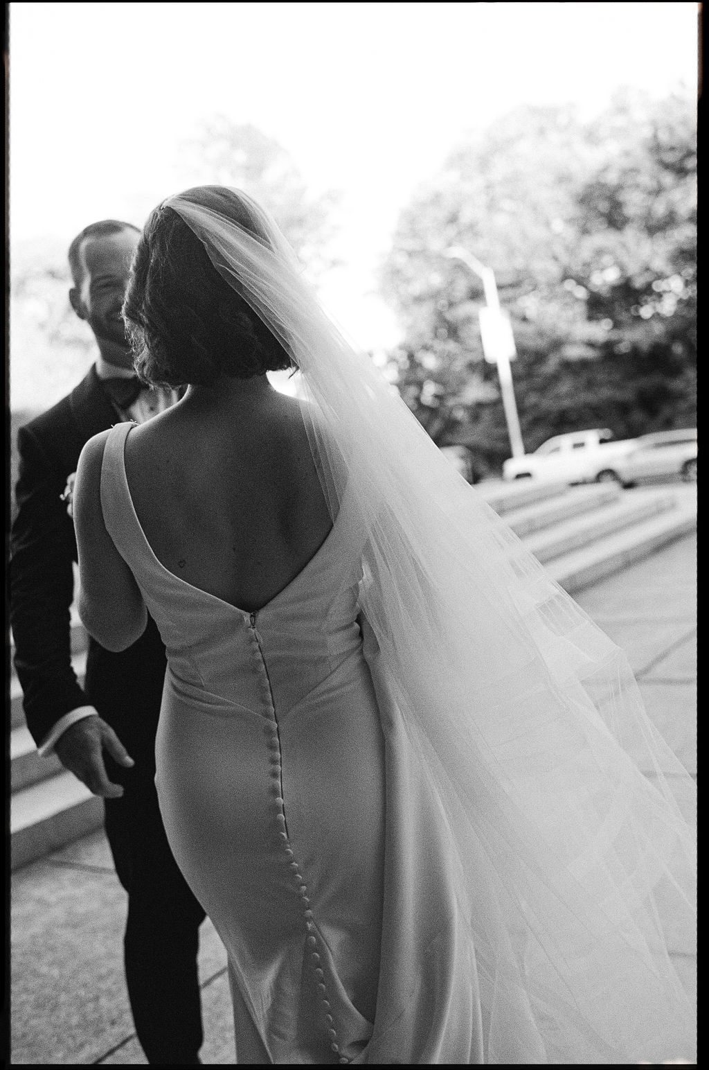A bride in a classic white wedding dress with button details on the back and flowing veil shown from behind.