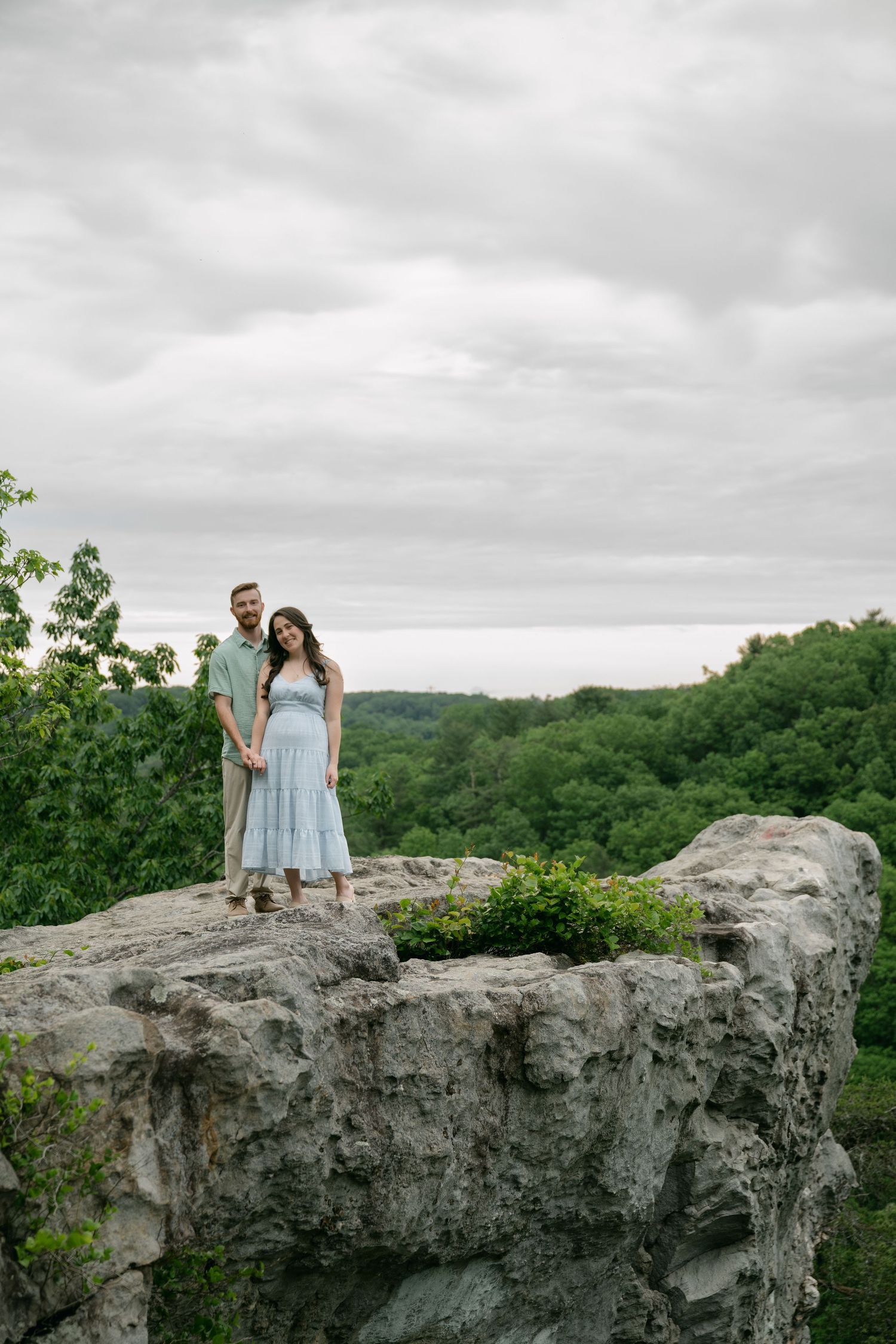 Couple standing on a rocky cliff overlooking a vast green forest under a cloudy sky.