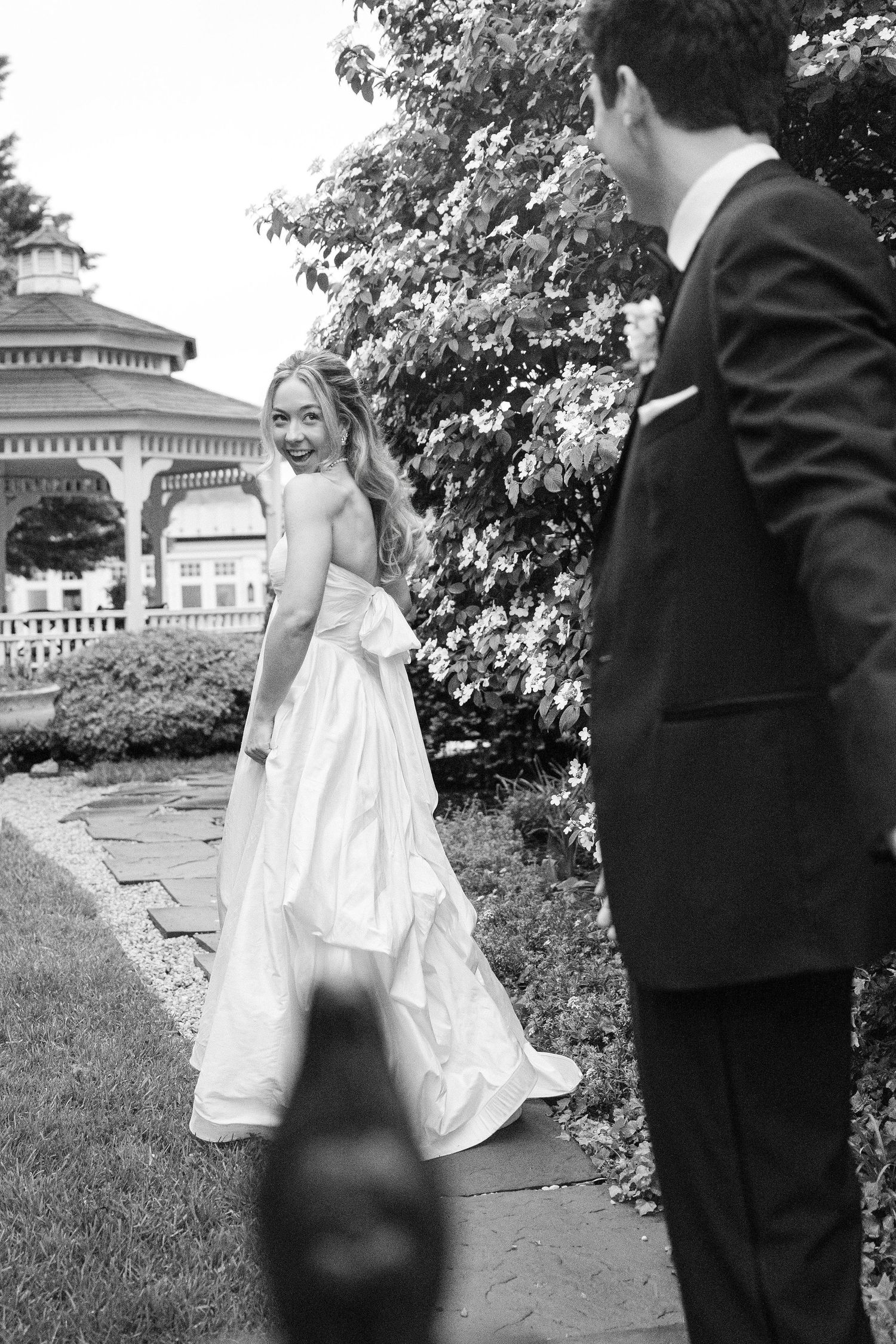 Bride in white gown glances back at groom in black tuxedo on garden path near gazebo in black and white wedding photo.