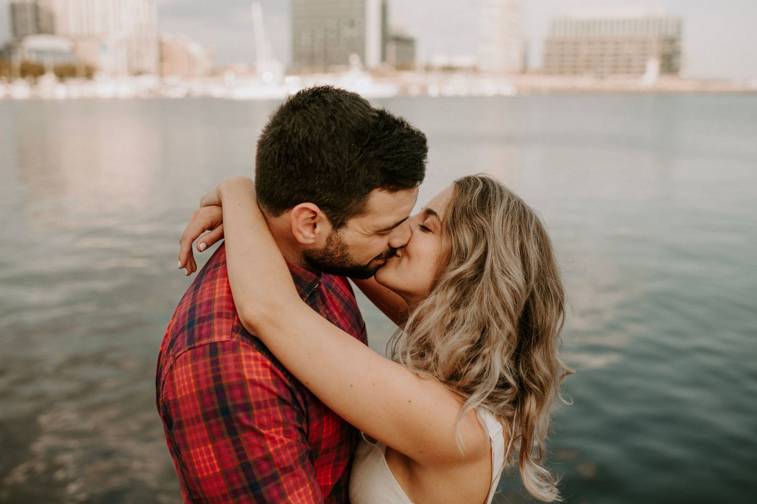 Intimate couple sharing a kiss by the water with urban backdrop.