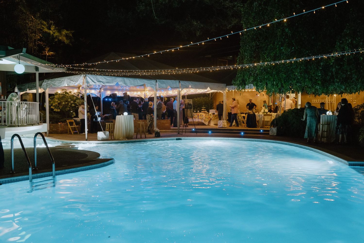 Nighttime poolside party setup with string lights and tents creating an elegant outdoor atmosphere.