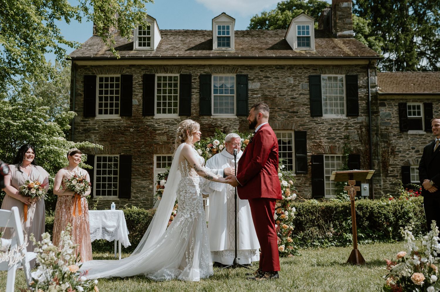 Wedding ceremony in progress on lawn of historic stone house with white chairs and floral decorations.