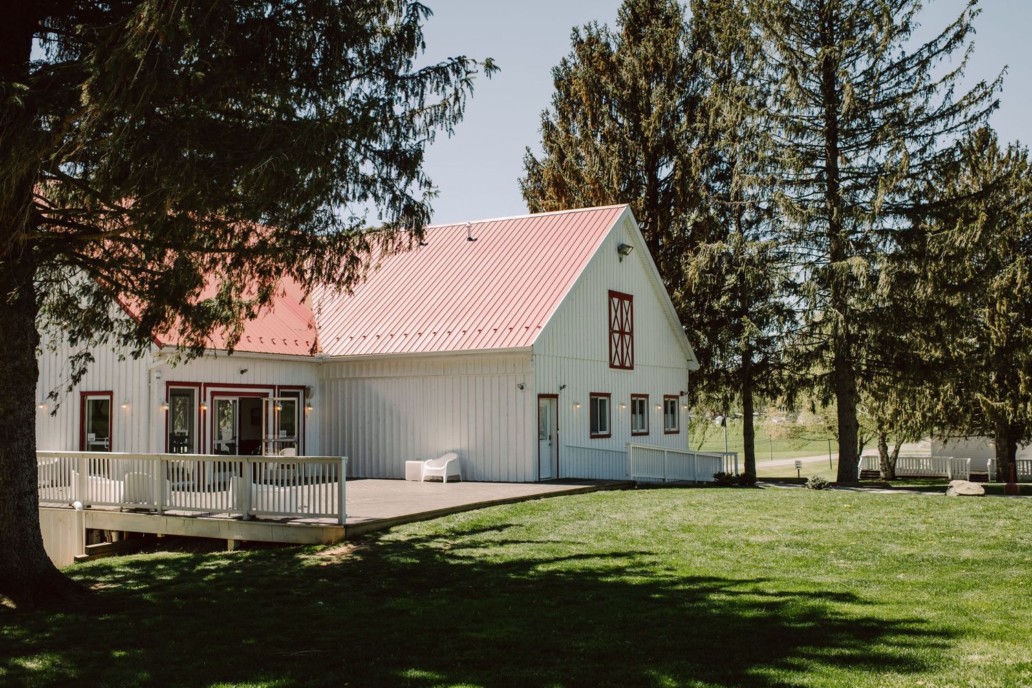 White farmhouse with red roof and wrap-around porch among pine trees.