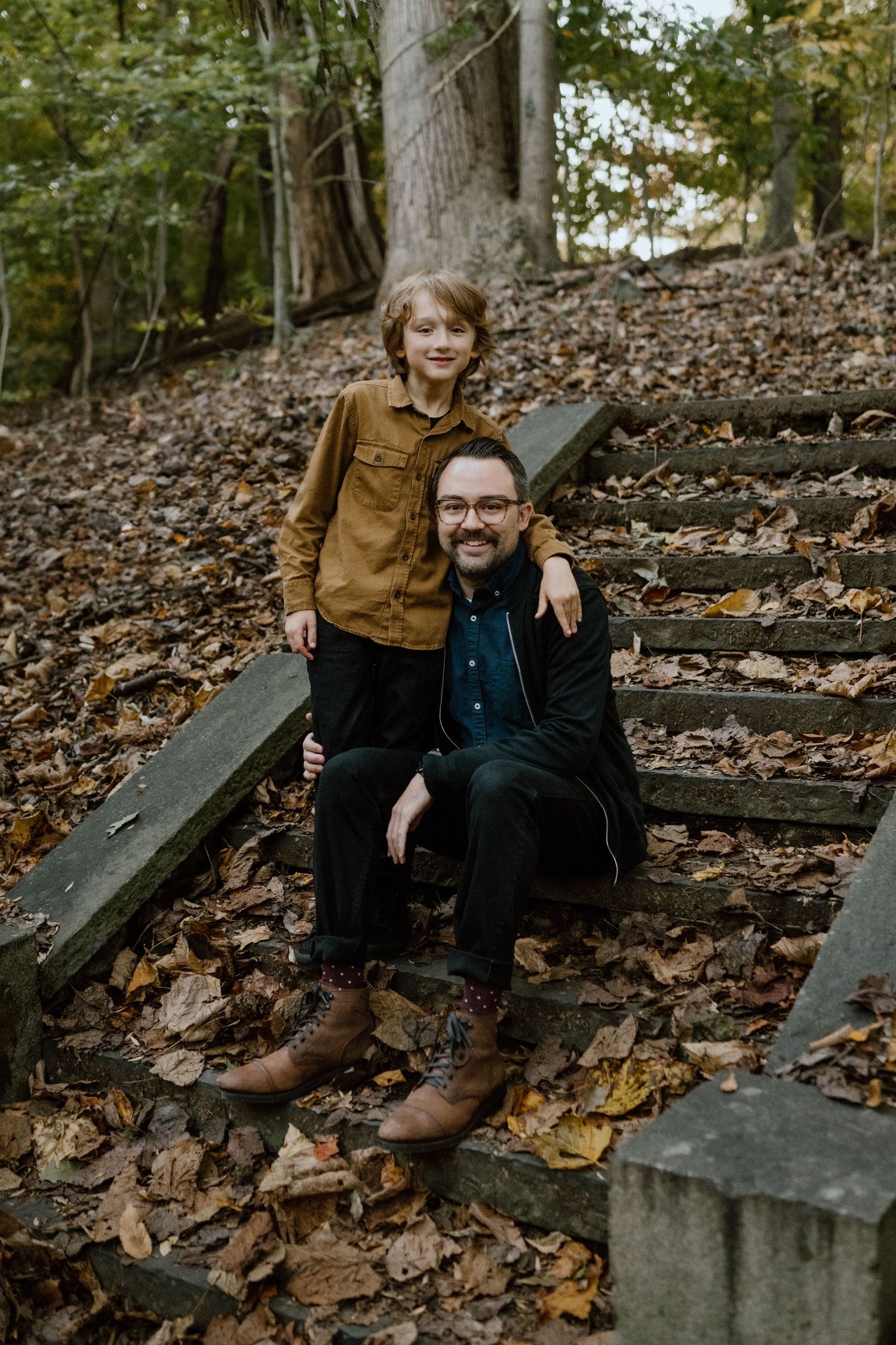 Two people pose together on wooden steps covered in fallen autumn leaves in a wooded setting.