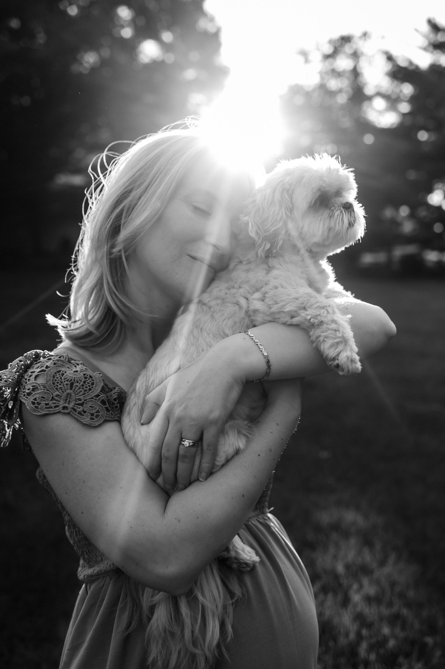Black and white portrait of a woman holding a small white dog with sunlight behind them.
