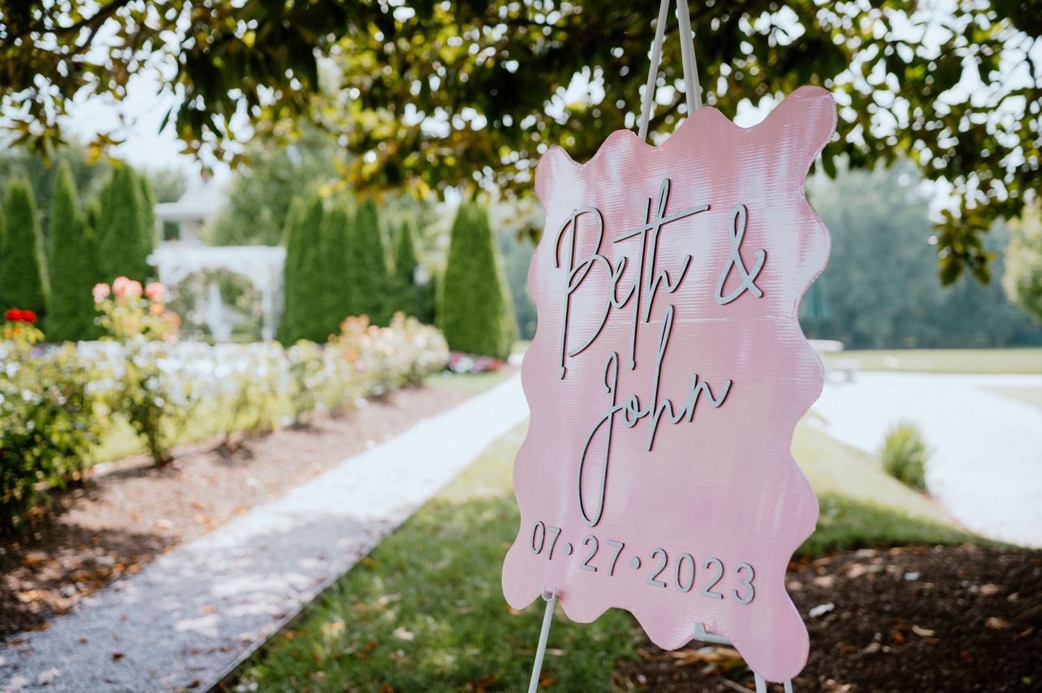 A pink wedding sign with names and date displayed in an outdoor garden setting.