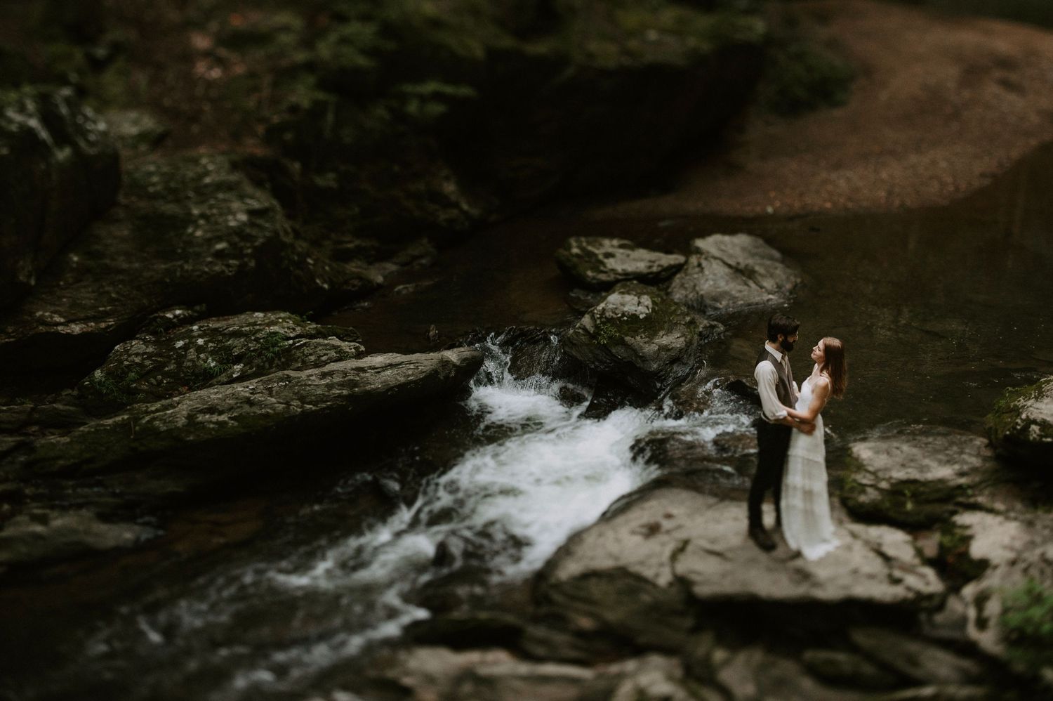 Wedding couple stands on dramatic rocky overlook above rushing forest waterfall.