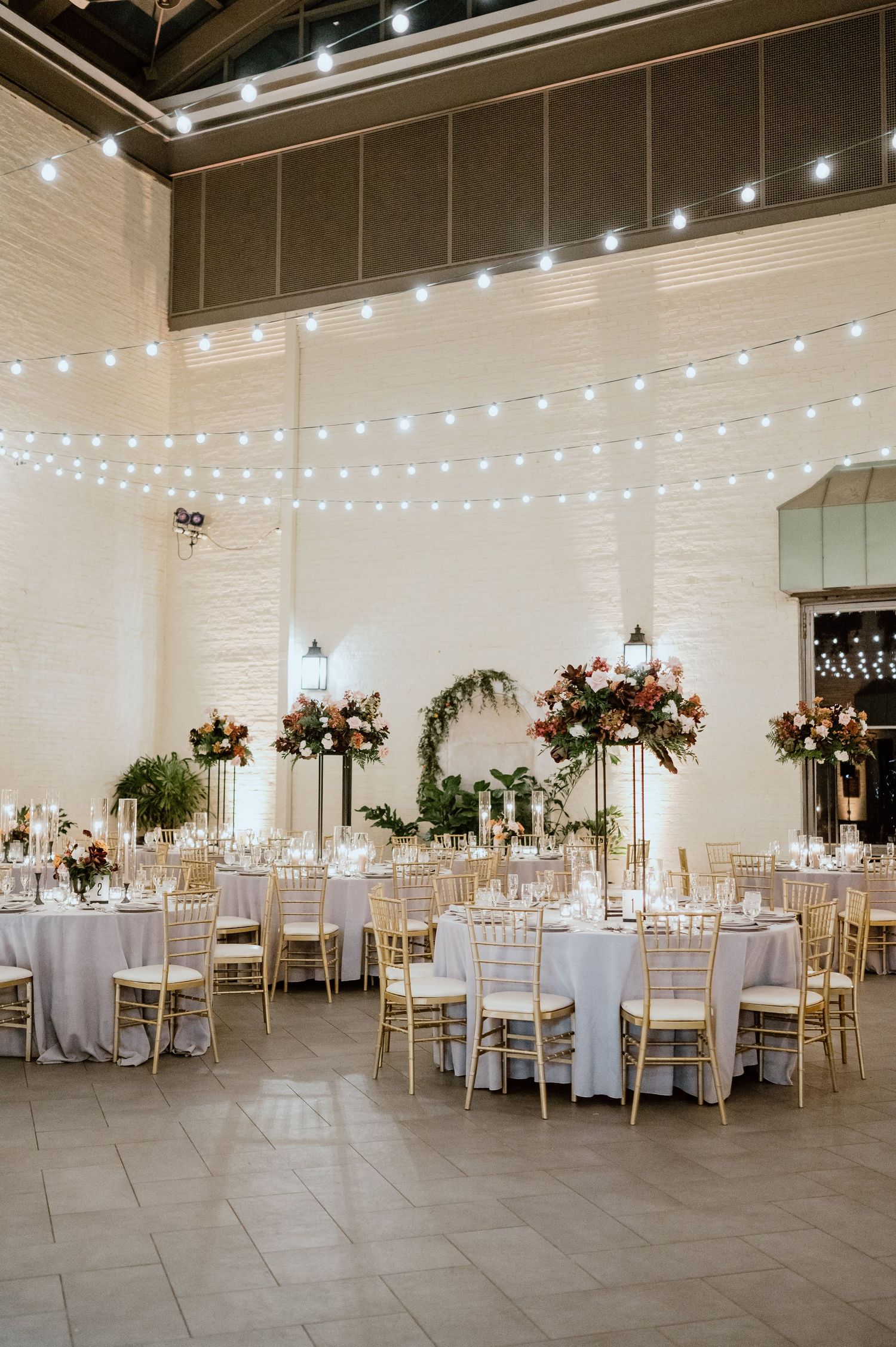 Elegant wedding reception hall with string lights and floral centerpieces on round tables with gold chairs.
