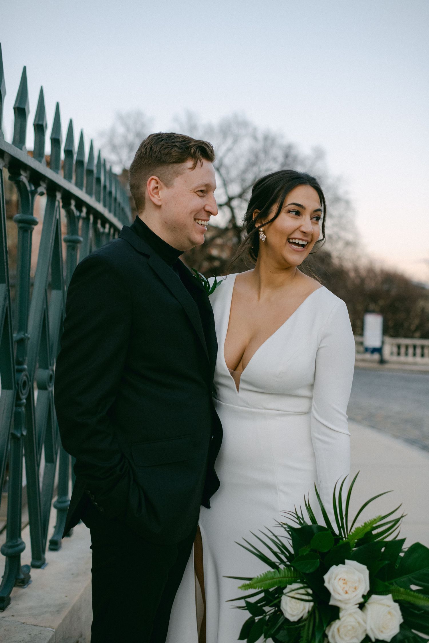 Modern wedding couple posing by iron fence with white floral bouquet at dusk.