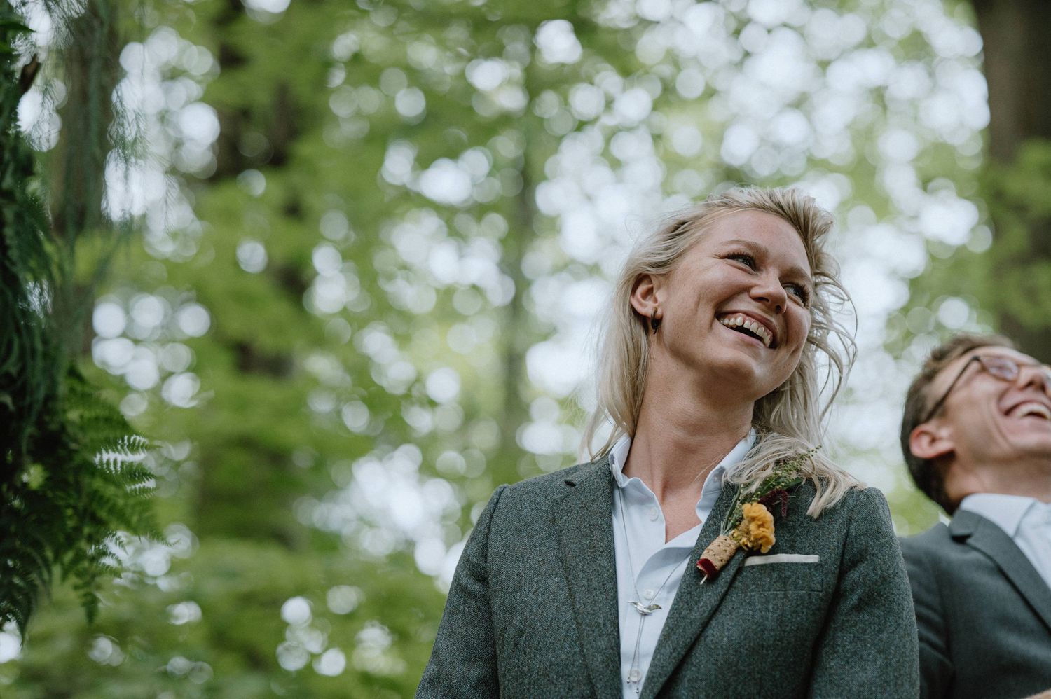 Wedding participants share a genuine laugh in a sunlit forest setting.