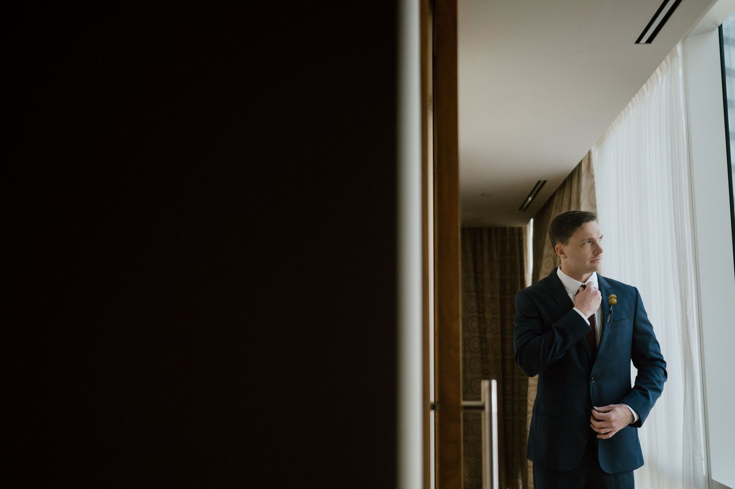 Dramatic side-lit portrait of person in suit adjusting tie by window.