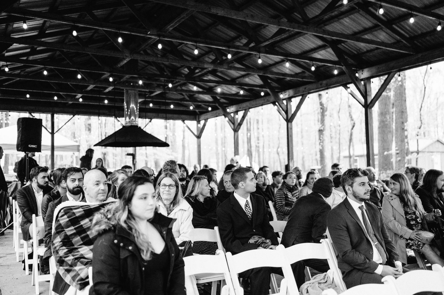Wedding guests seated under covered pavilion with string lights in black and white.