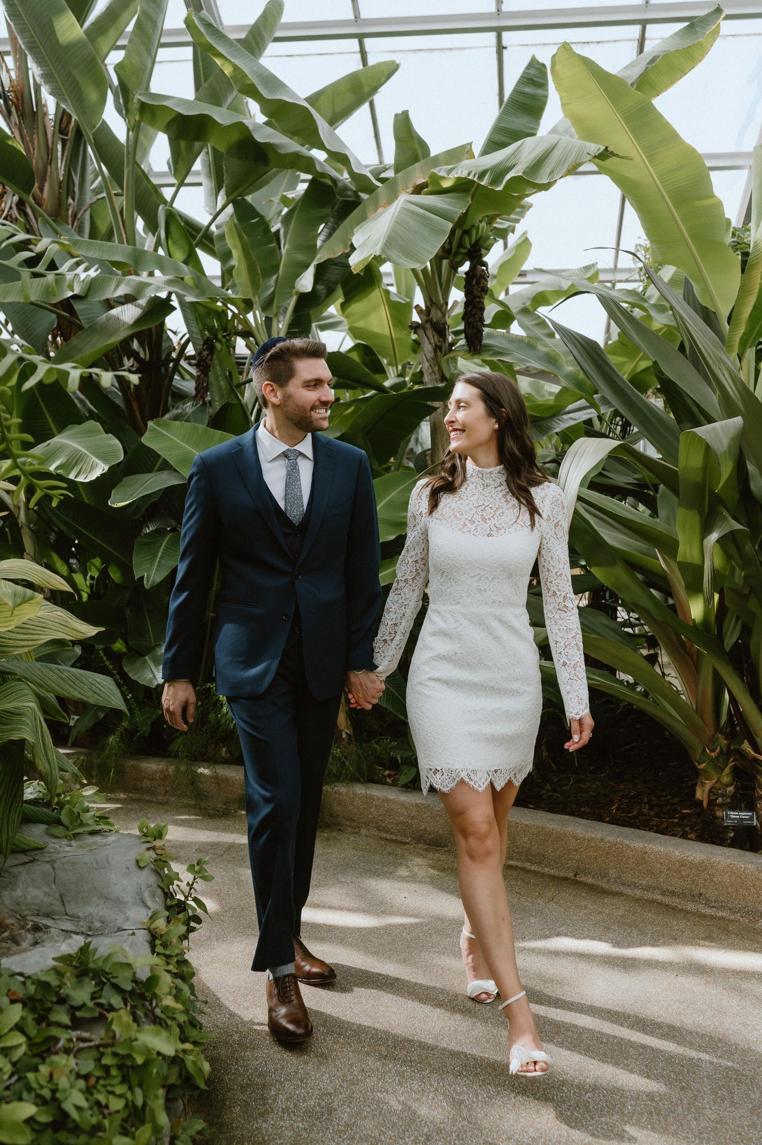 A couple strolling through a tropical greenhouse, she in white lace and he in a navy suit.