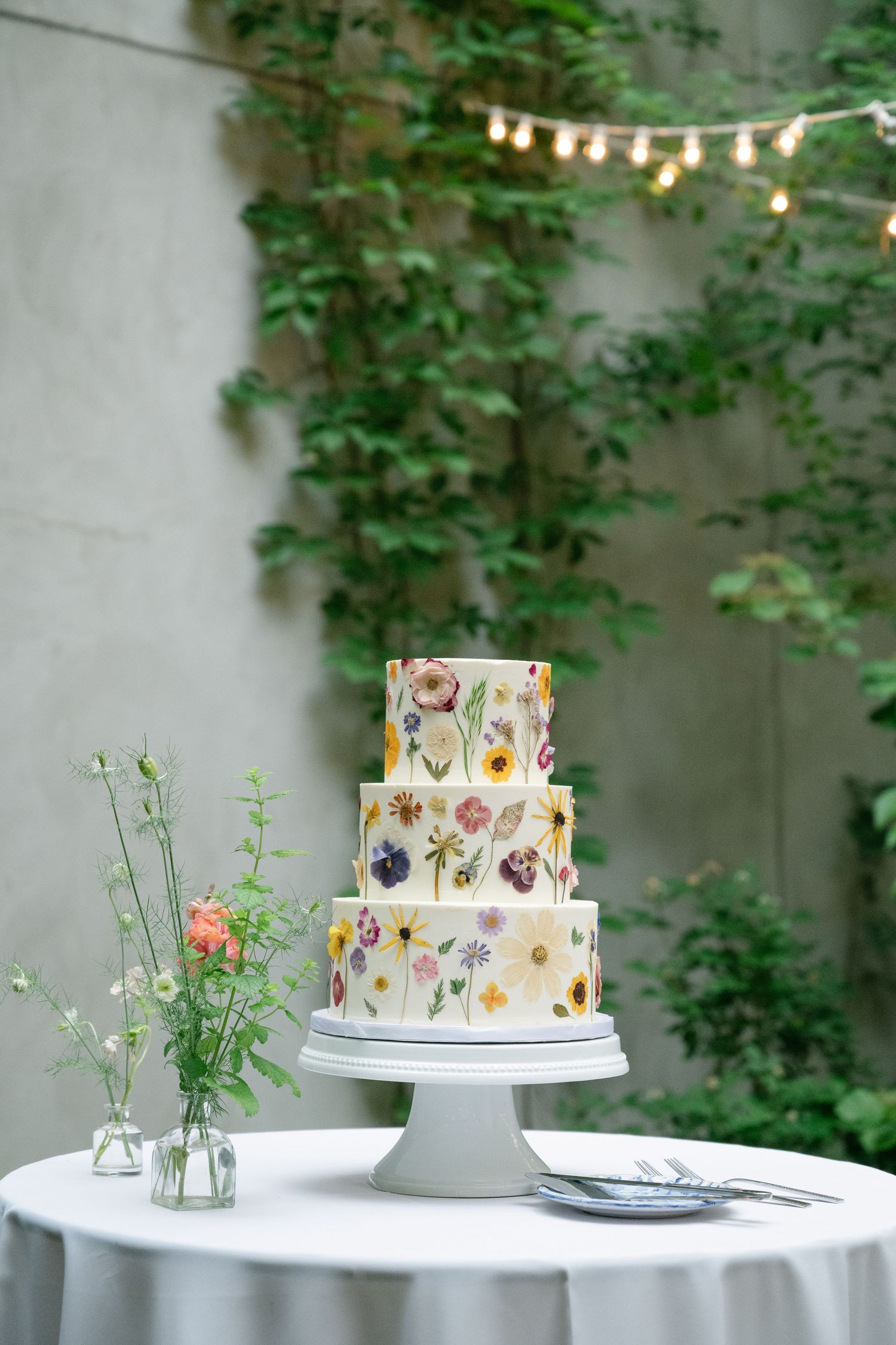 A three-tiered wedding cake decorated with pressed edible flowers sits on a white stand with string lights and greenery behind.