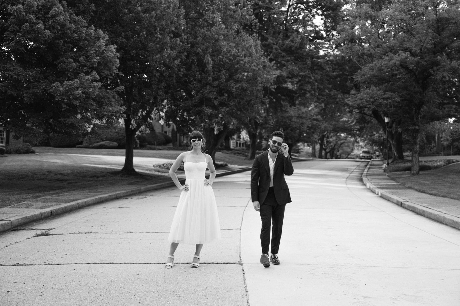 A black and white photo of a couple walking and dancing playfully on a tree-lined path.