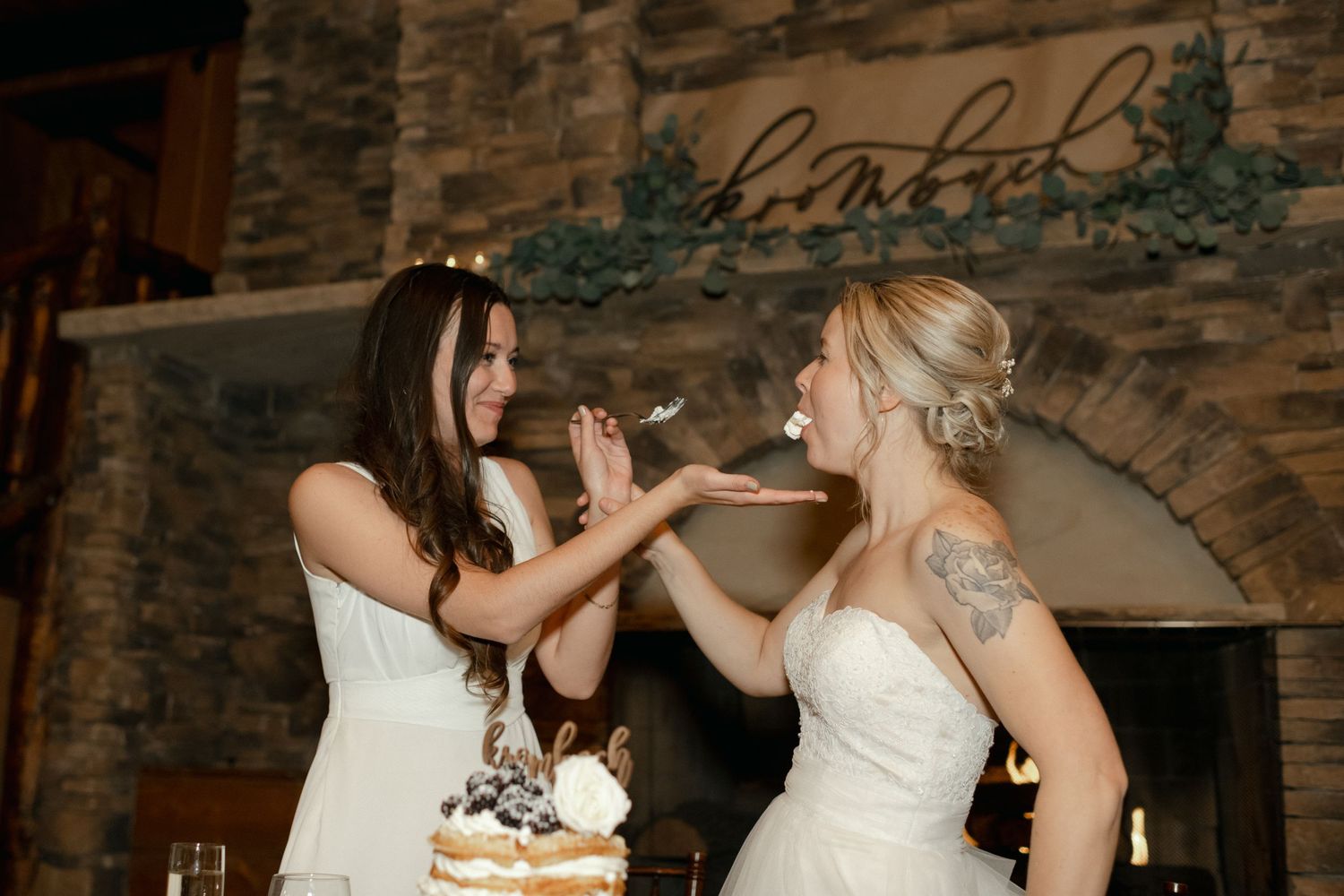 Two people in wedding attire sharing cake at a rustic stone fireplace reception.
