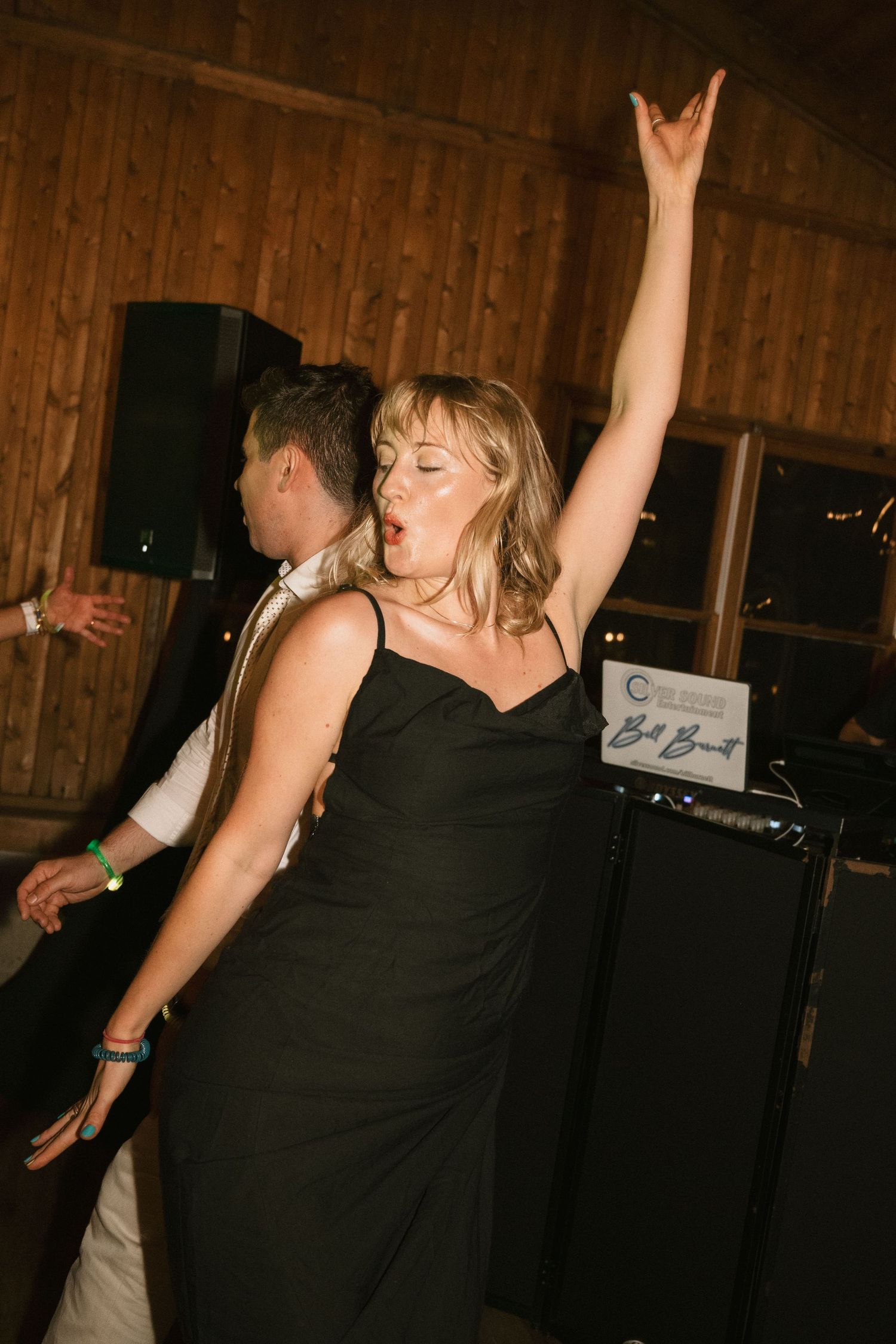 A dancer in a black dress shows enthusiasm while dancing at a party venue with speakers visible.