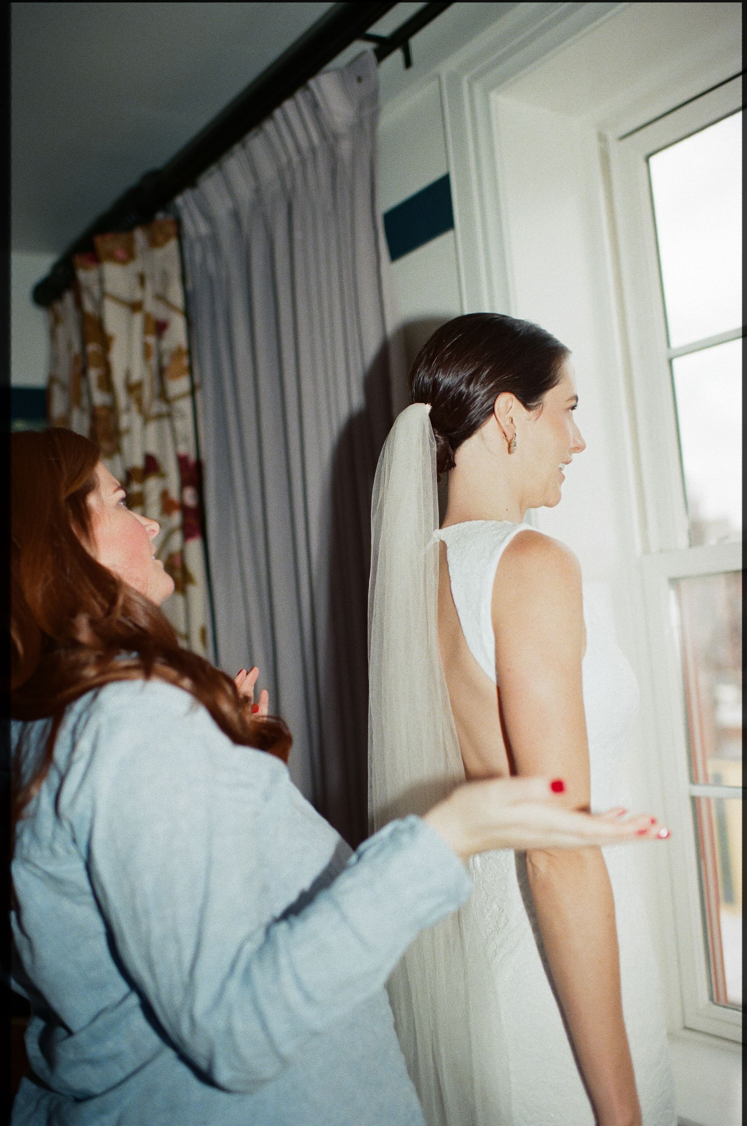 Bride in white dress getting final adjustments near a window during wedding preparations.