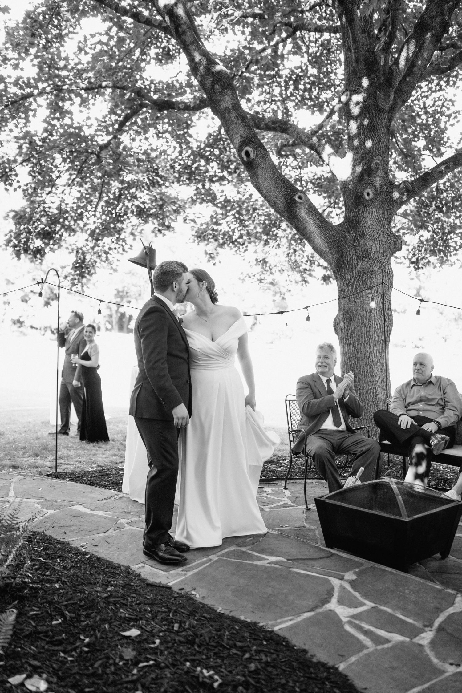 Wedding couple shares intimate moment under large tree with string lights at outdoor reception.