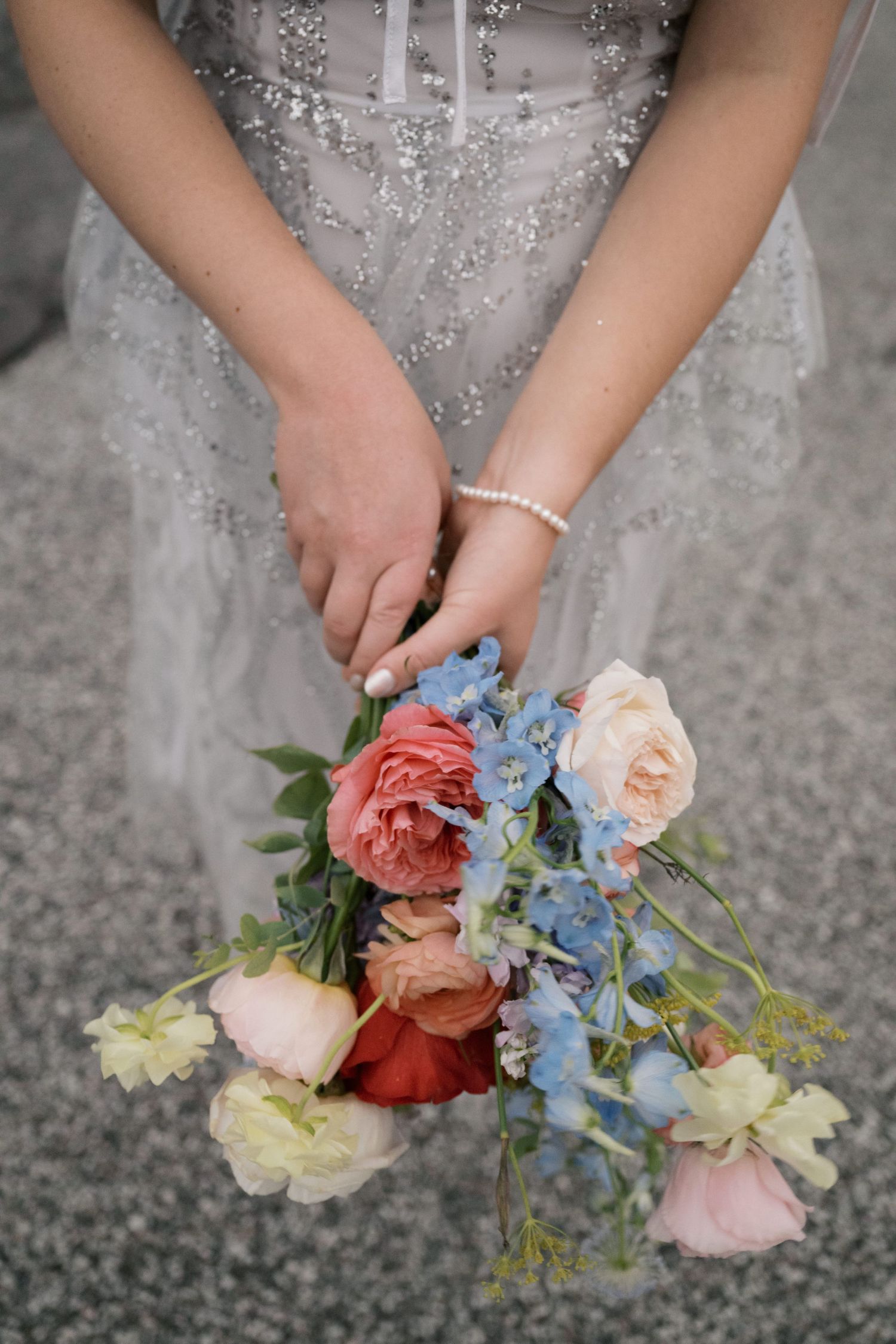 Close-up of hands holding a delicate bouquet with coral and blue flowers and a pearl bracelet.