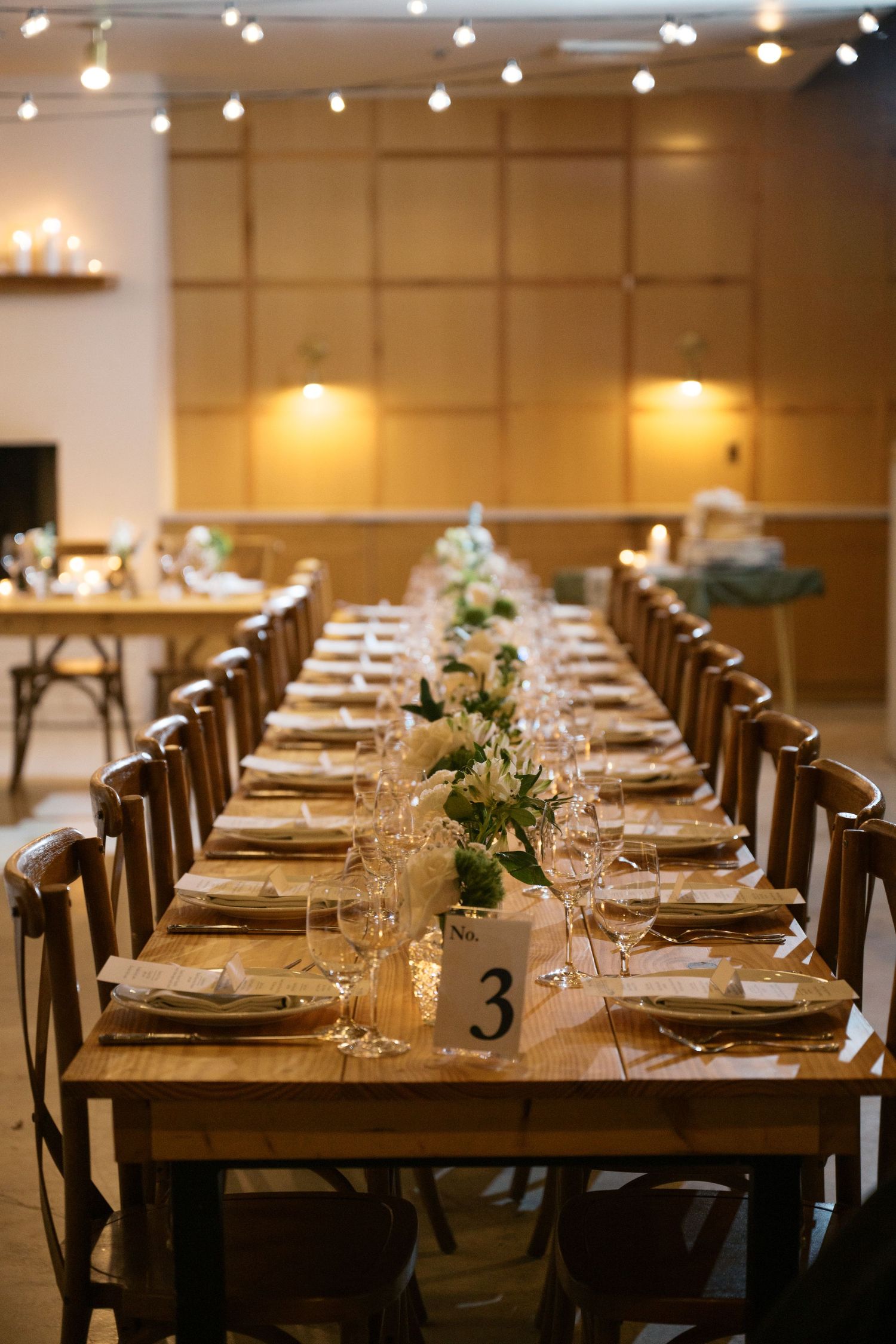 Long wooden dining table set for a wedding reception with white floral centerpieces and string lights overhead.
