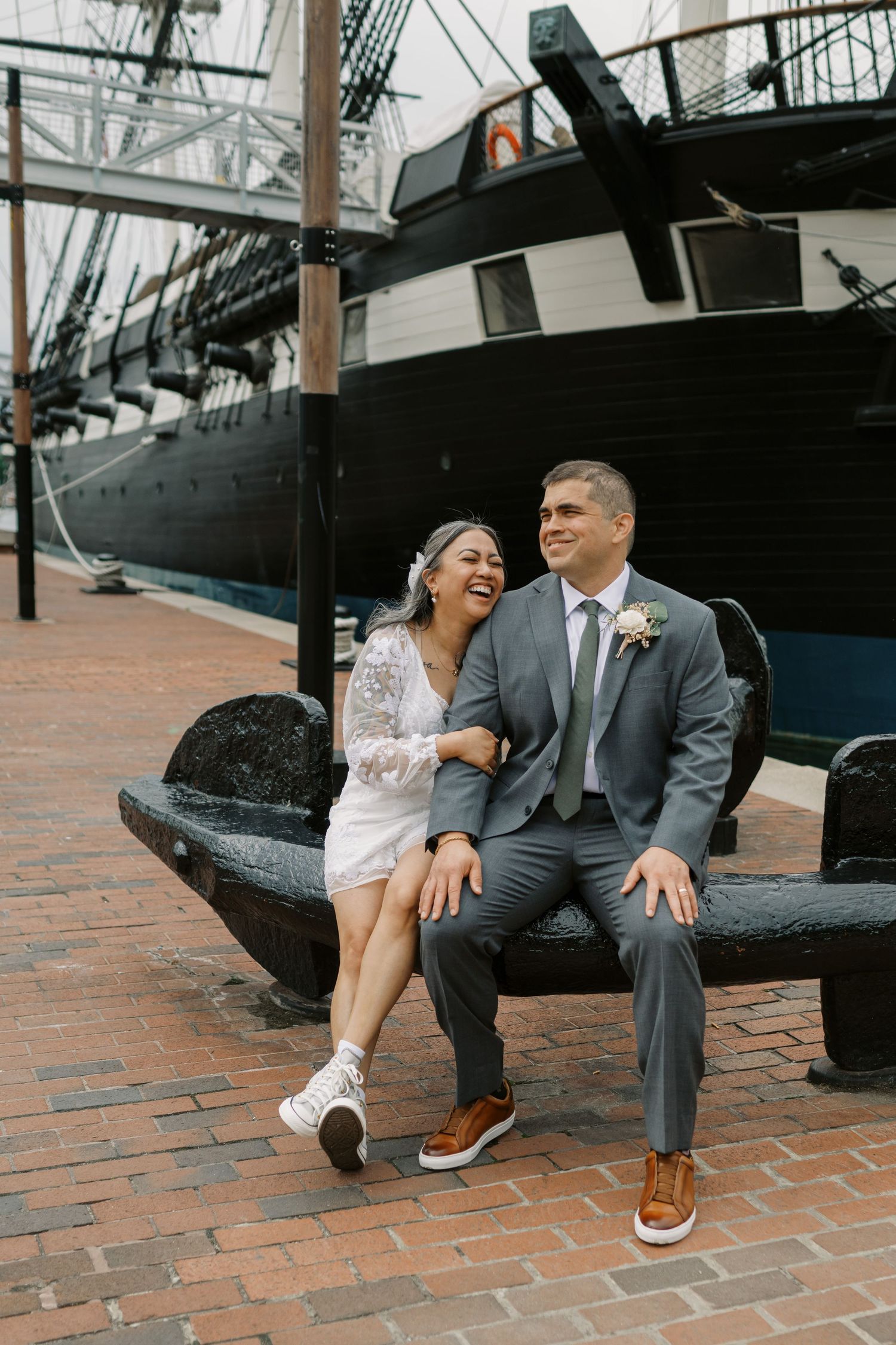A couple shares a laugh while sitting on an anchor near a historic ship.
