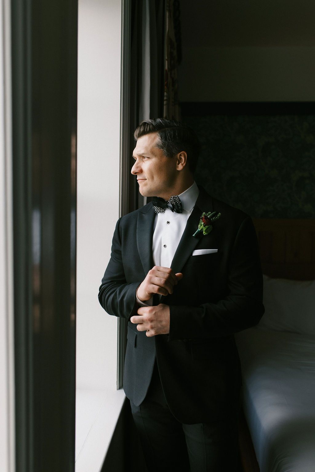 Groom in classic black tuxedo and bow tie adjusts his cufflinks in moody window light.
