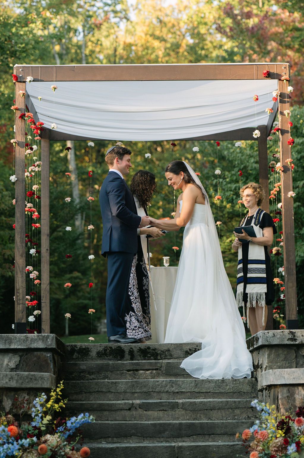 Wedding ceremony under a wooden canopy with white fabric and floral decorations.